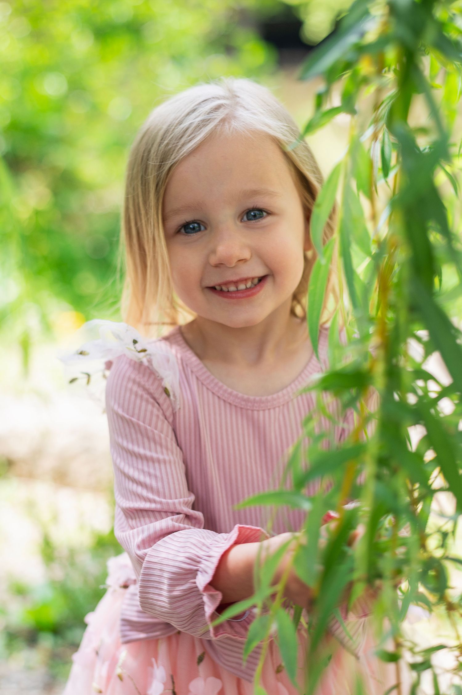 Toddler girl smiling through green branches during summer milestone session at Gahanna Creekside.
