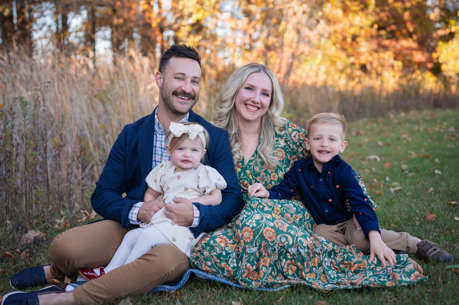 Family of four sitting on a blanket during a fall mini session, surrounded by golden light and tall autumn grasses.