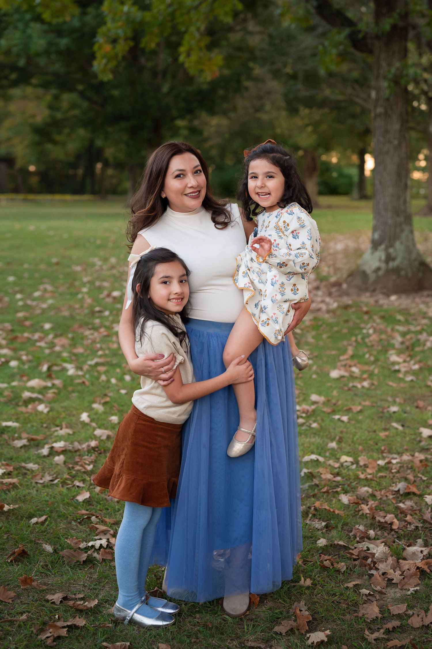Mom in a blue tulle skirt cuddles both daughters during a playful fall family photo session in a central Ohio park.