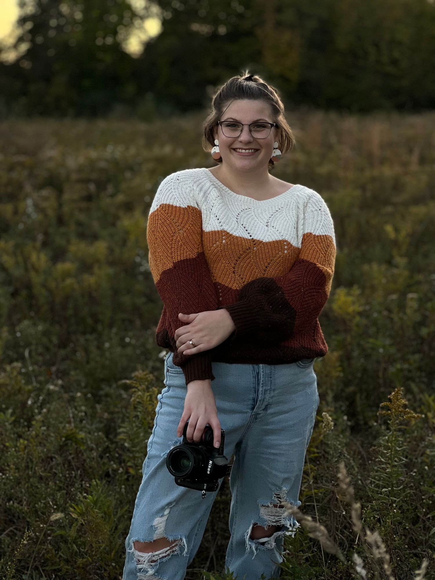 Photographer portrait in fall field, Smiling woman in a cozy striped sweater and ripped jeans holds a Nikon camera while standing in a field