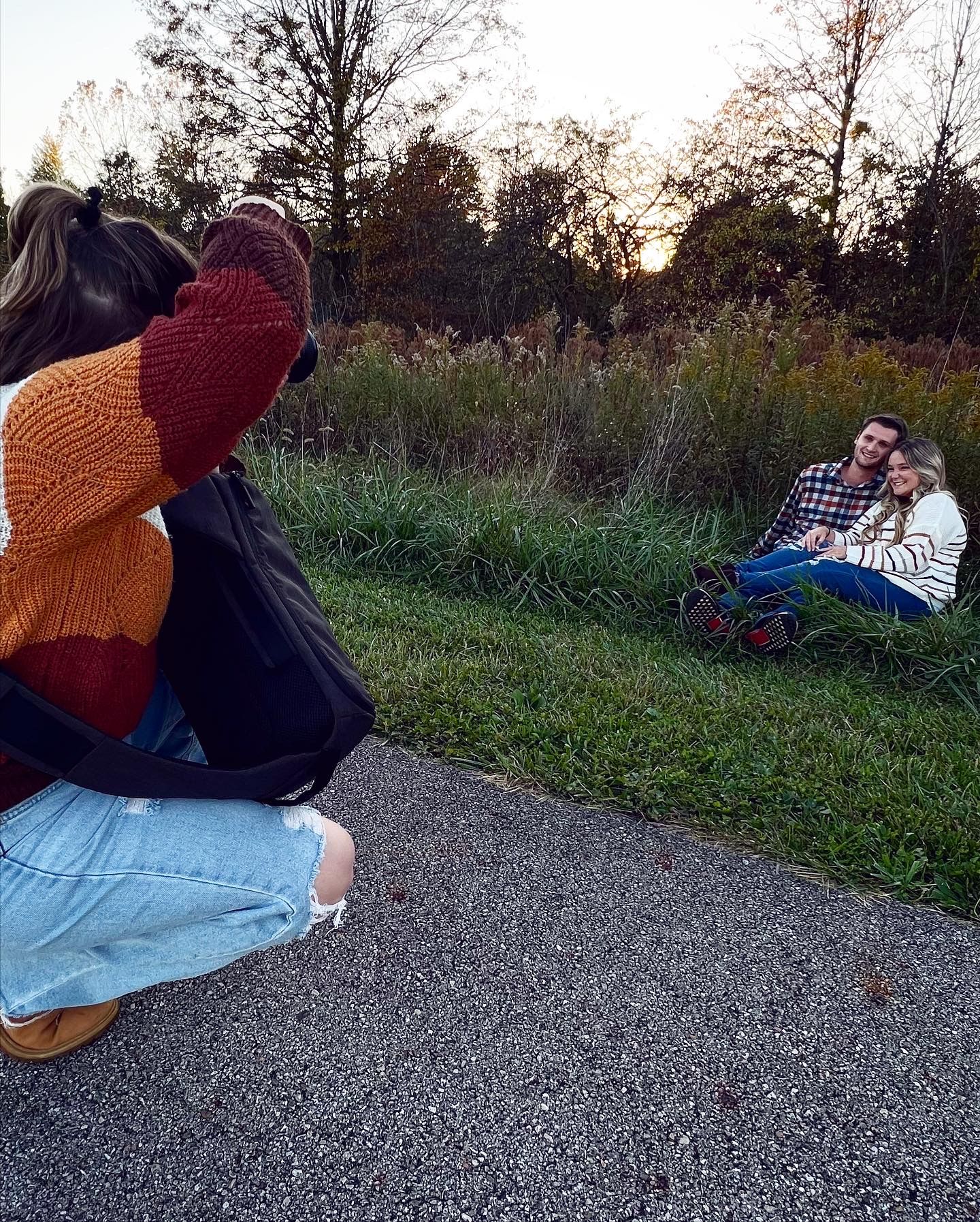 Behind-the-scenes fall photo session, Photographer kneels on a paved path, wearing a striped fall sweater and ripped jeans.