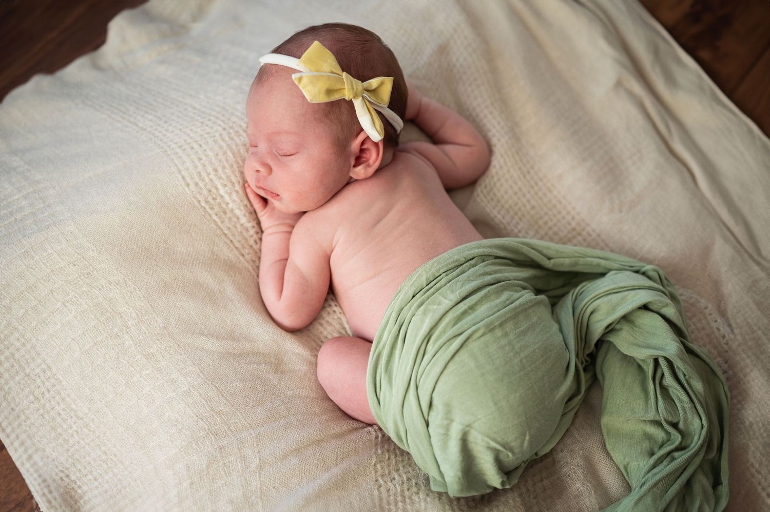Sleeping newborn girl posed on a blanket with a yellow bow headband and wrapped in a green swaddle.