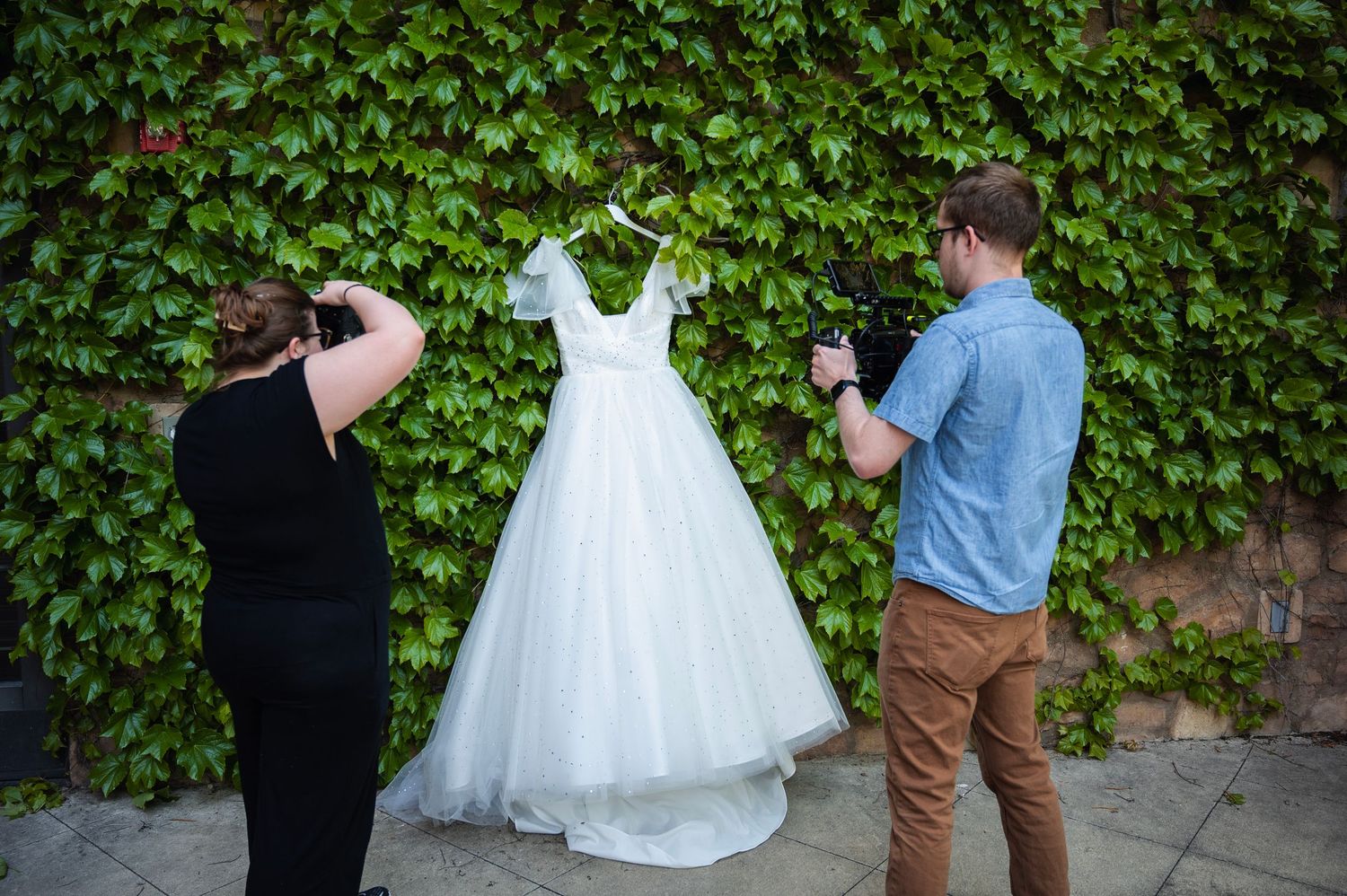 Behind the scenes of a wedding dress photo setup with photographer and videographer at a lush green ivy wall.