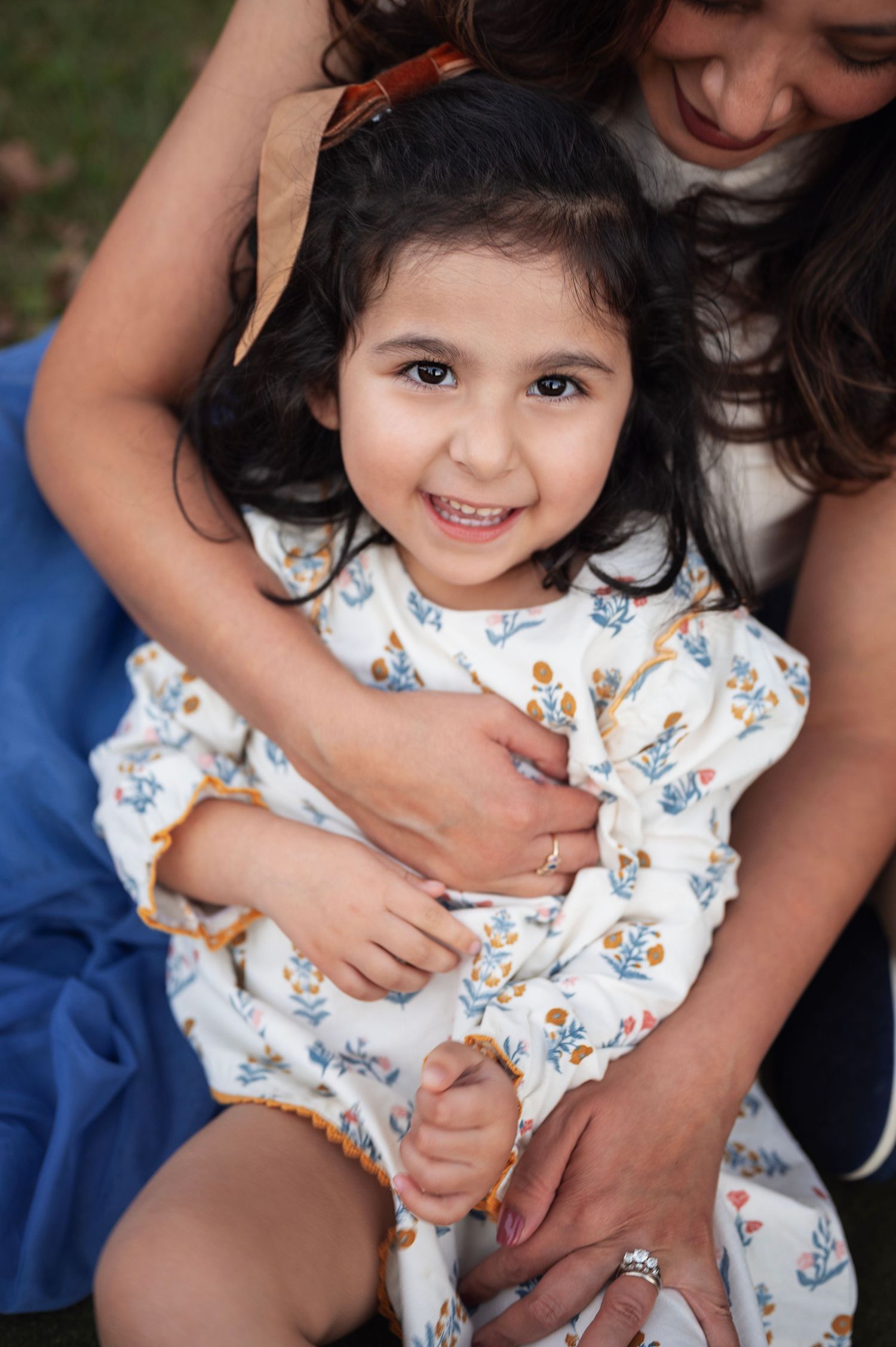 Smiling toddler in a floral dress cuddles in her mother’s arms, showing off her brown bow and long lashes during fall family photos.