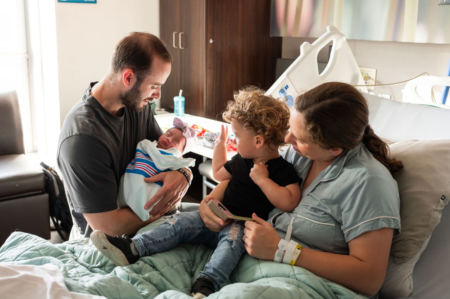 Family of four on hospital bed as big brother meets his baby sister for the first time, smiles and curiosity overflowing.