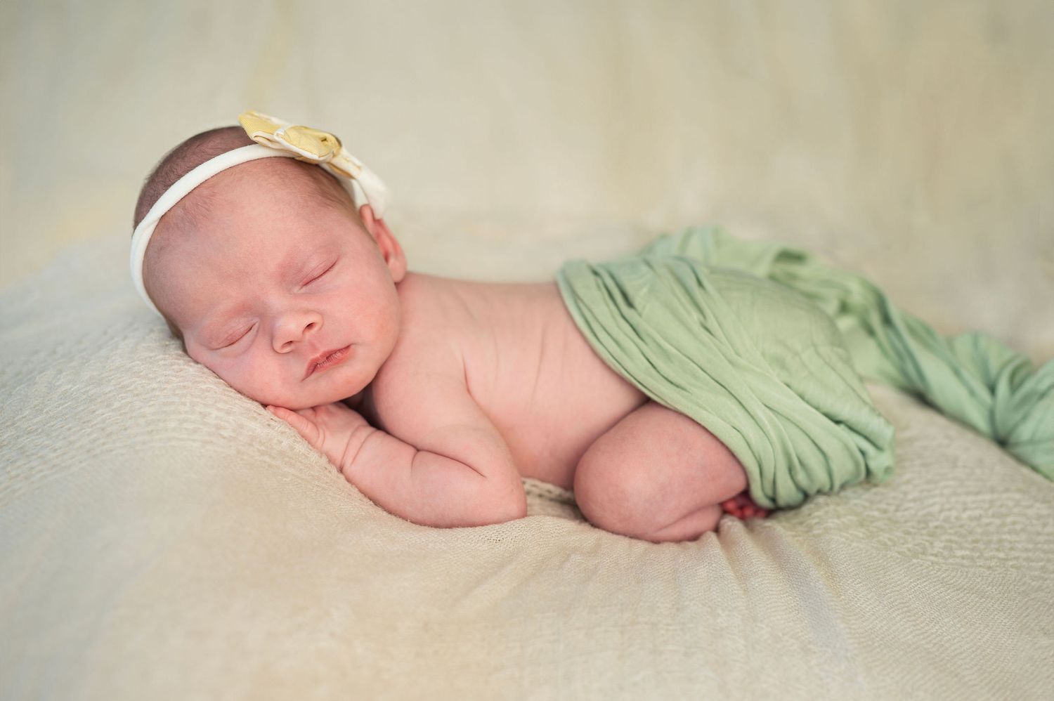 Newborn girl sleeps soundly on a soft blanket, swaddled in green with a yellow bow in this tender, cozy studio pose.