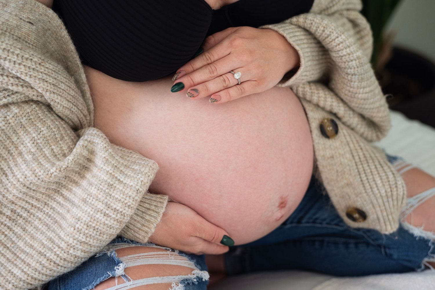 Pregnant woman in ripped jeans and cozy cardigan cradling her bare belly with manicured hands.