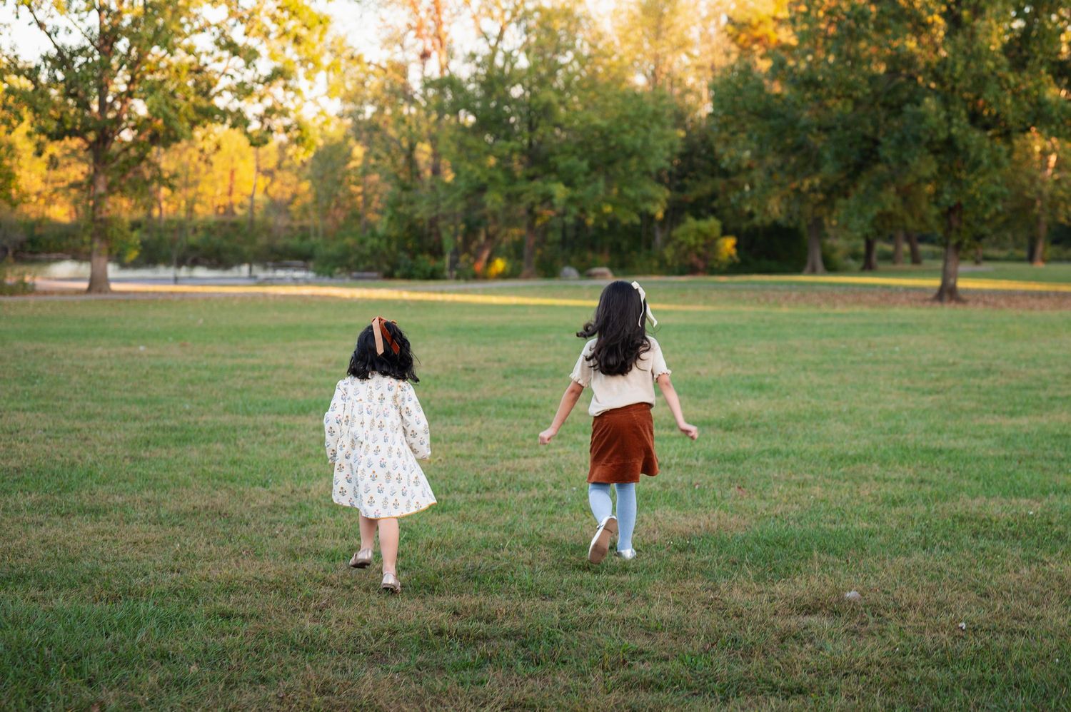 Two young sisters run through a park in coordinated outfits with matching hair bows as playful accessories during a family photo session.