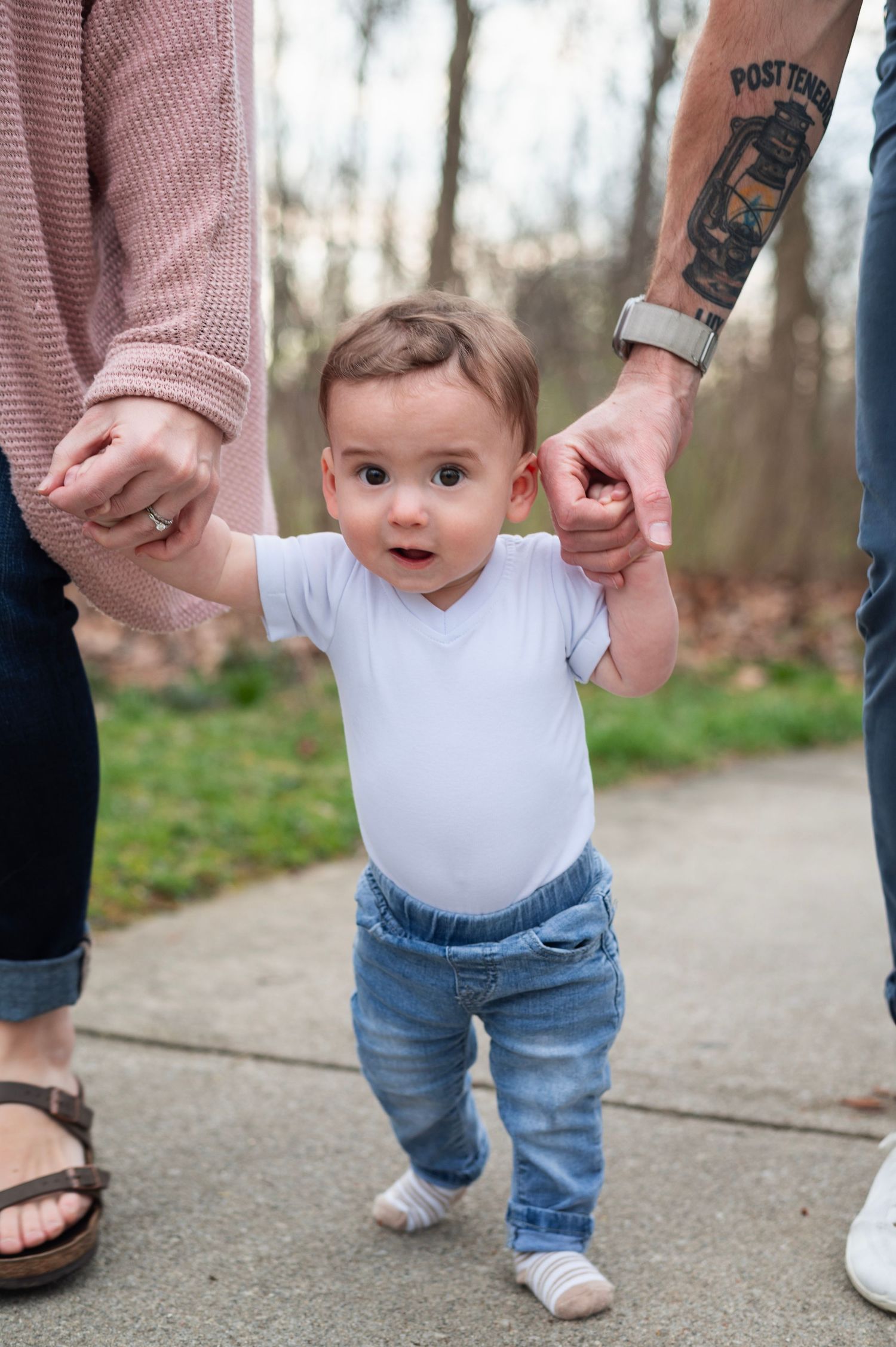 Baby takes first steps on a sidewalk, holding hands with mom and tattooed dad during a milestone session.