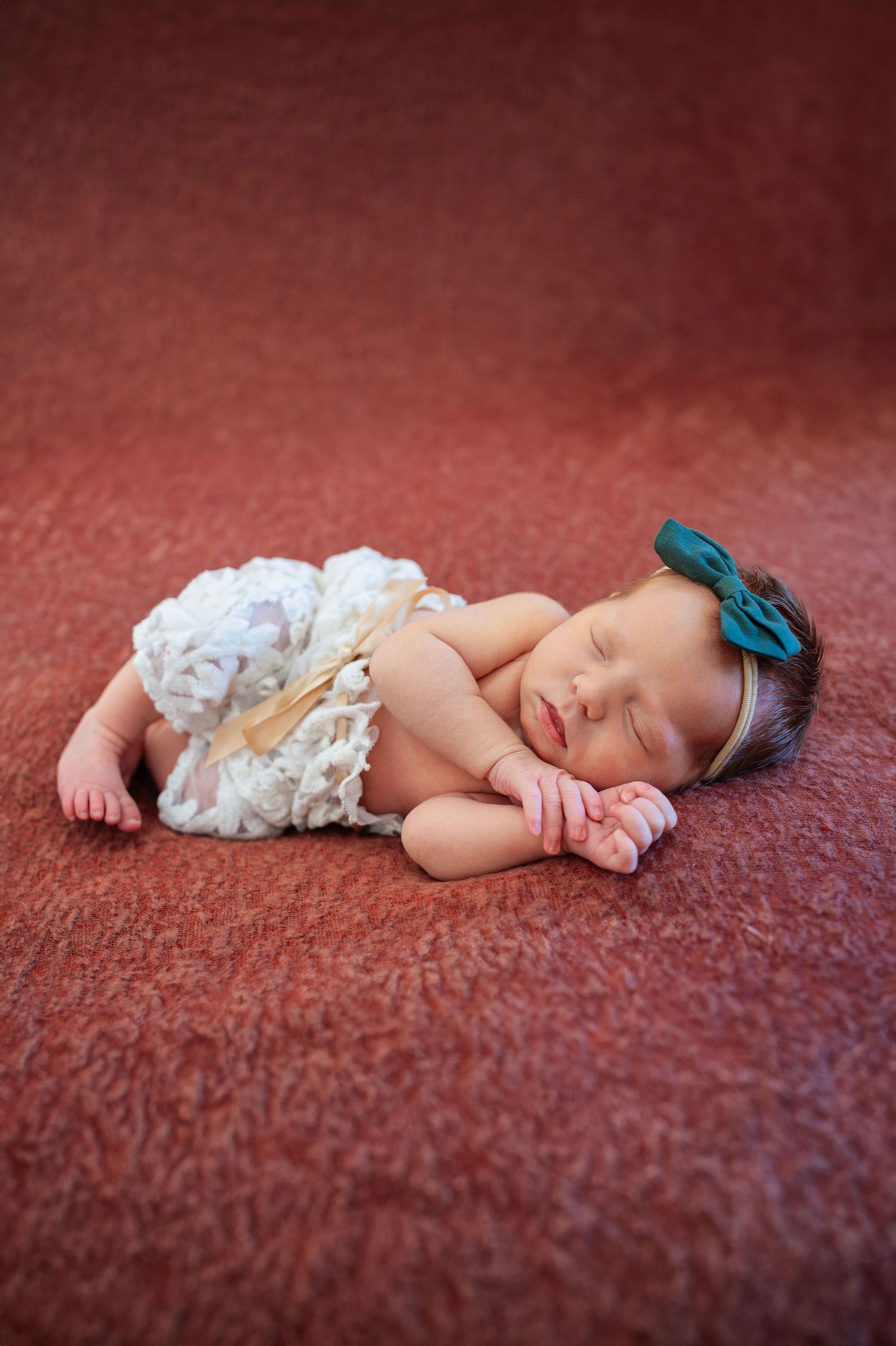 Newborn girl in lace pants and green bow sleeping on textured red blanket, posed studio session in Columbus.