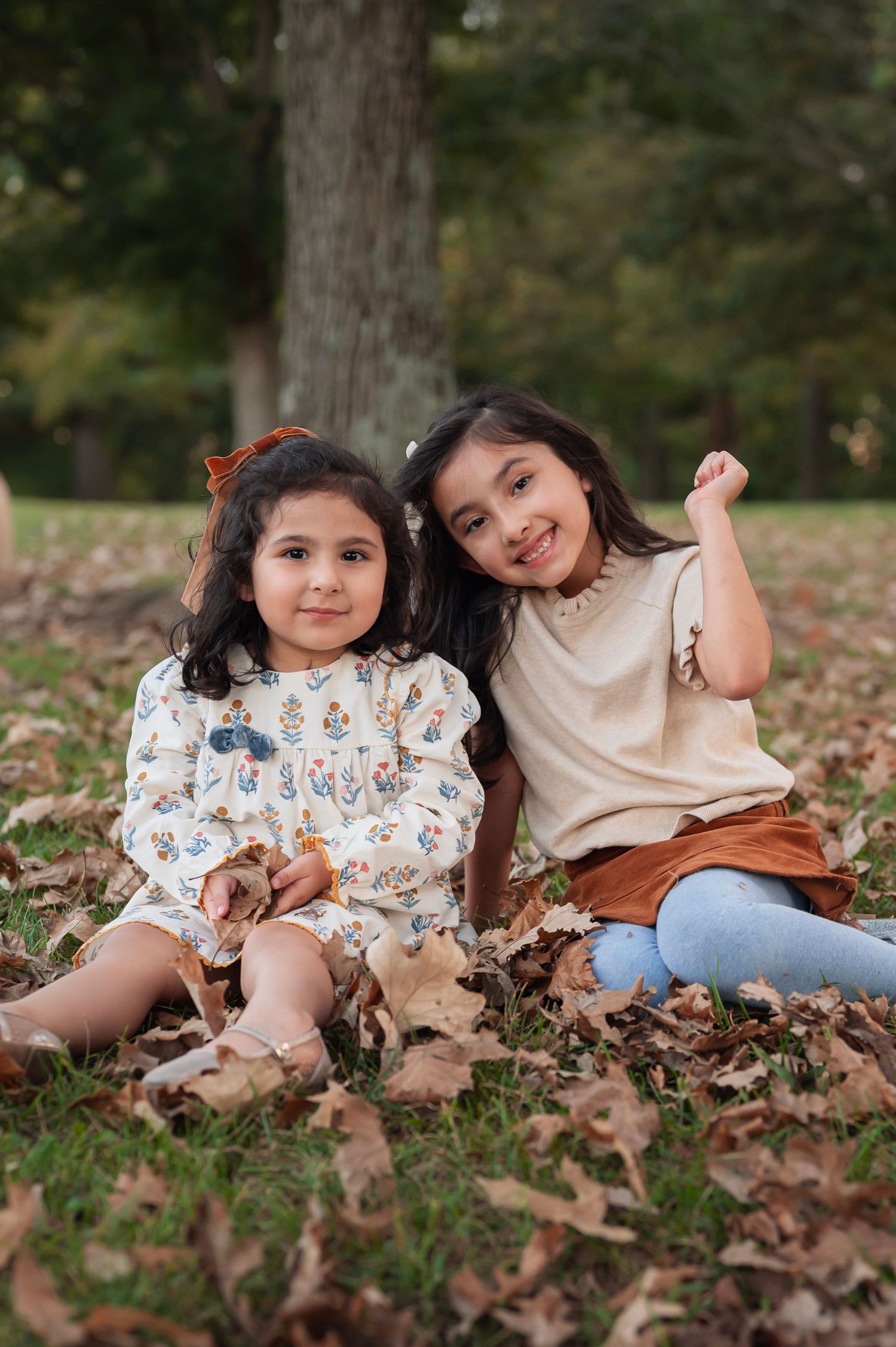 Sisters sit in a carpet of fallen leaves, smiling at the camera in cozy neutral outfits and hair bows for fall photos.