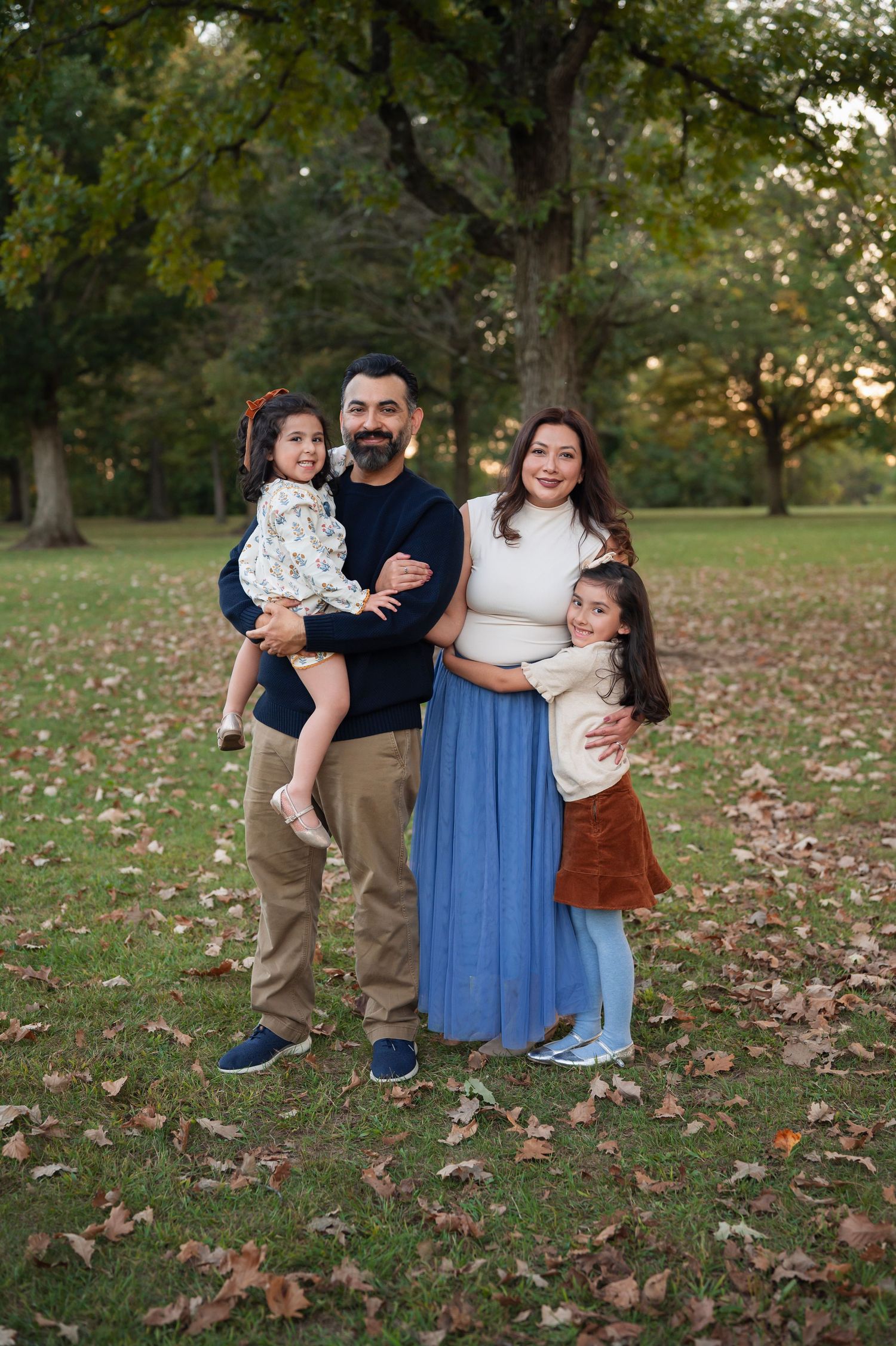 Westerville family photographer captures a cozy fall portrait of parents and two daughters standing together under tall trees at sunset.