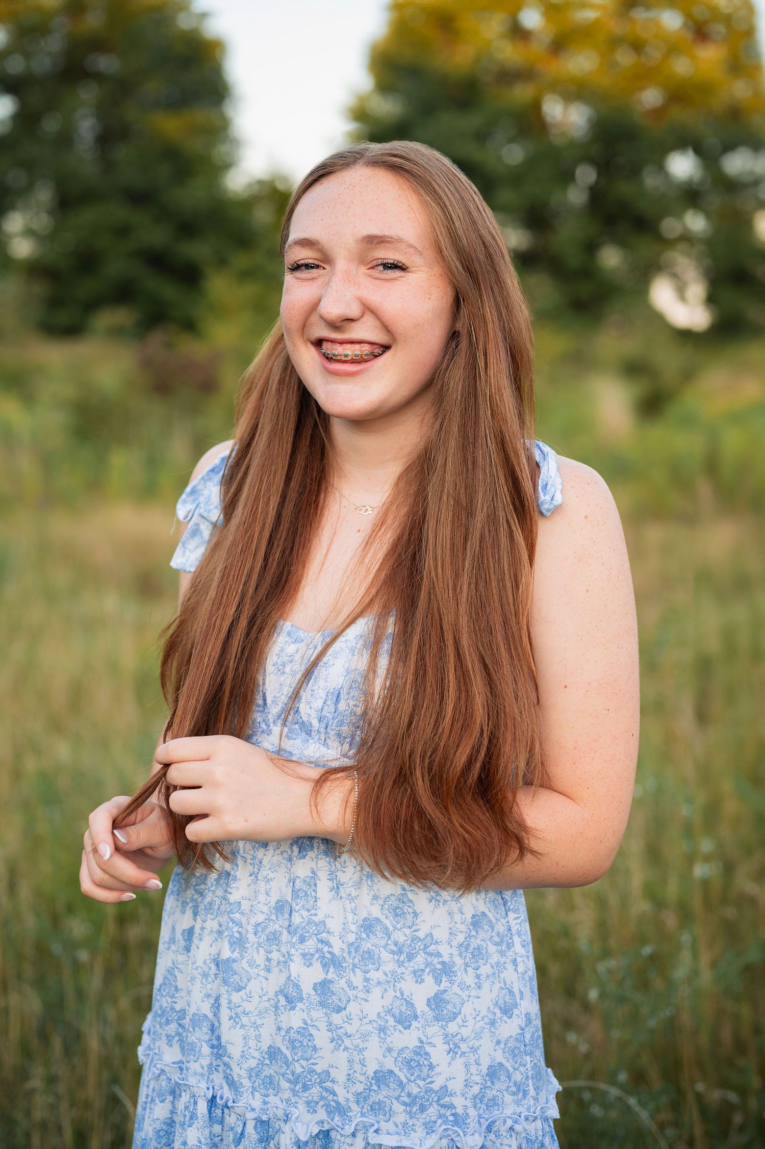 Senior girl with long red hair smiles brightly in a floral dress during a relaxed outdoor senior session.