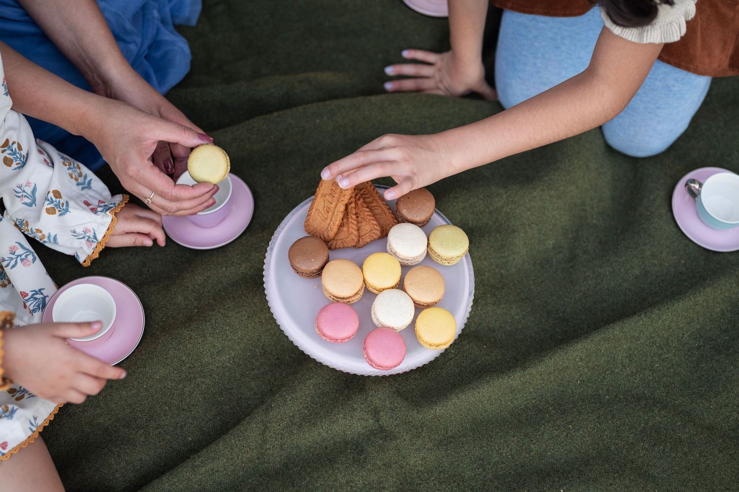 Hands reaching for colorful macarons and cookies on a cake stand during a family picnic tea party on a green blanket.