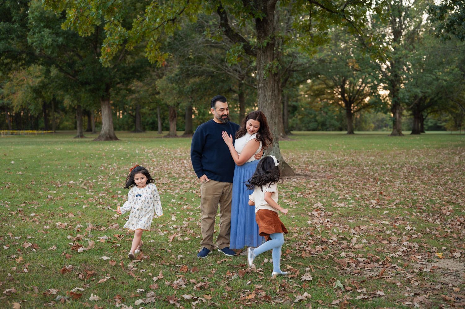 Parents stand laughing together on a leaf-covered lawn while their two daughters run circles around them in playful fall family photos.