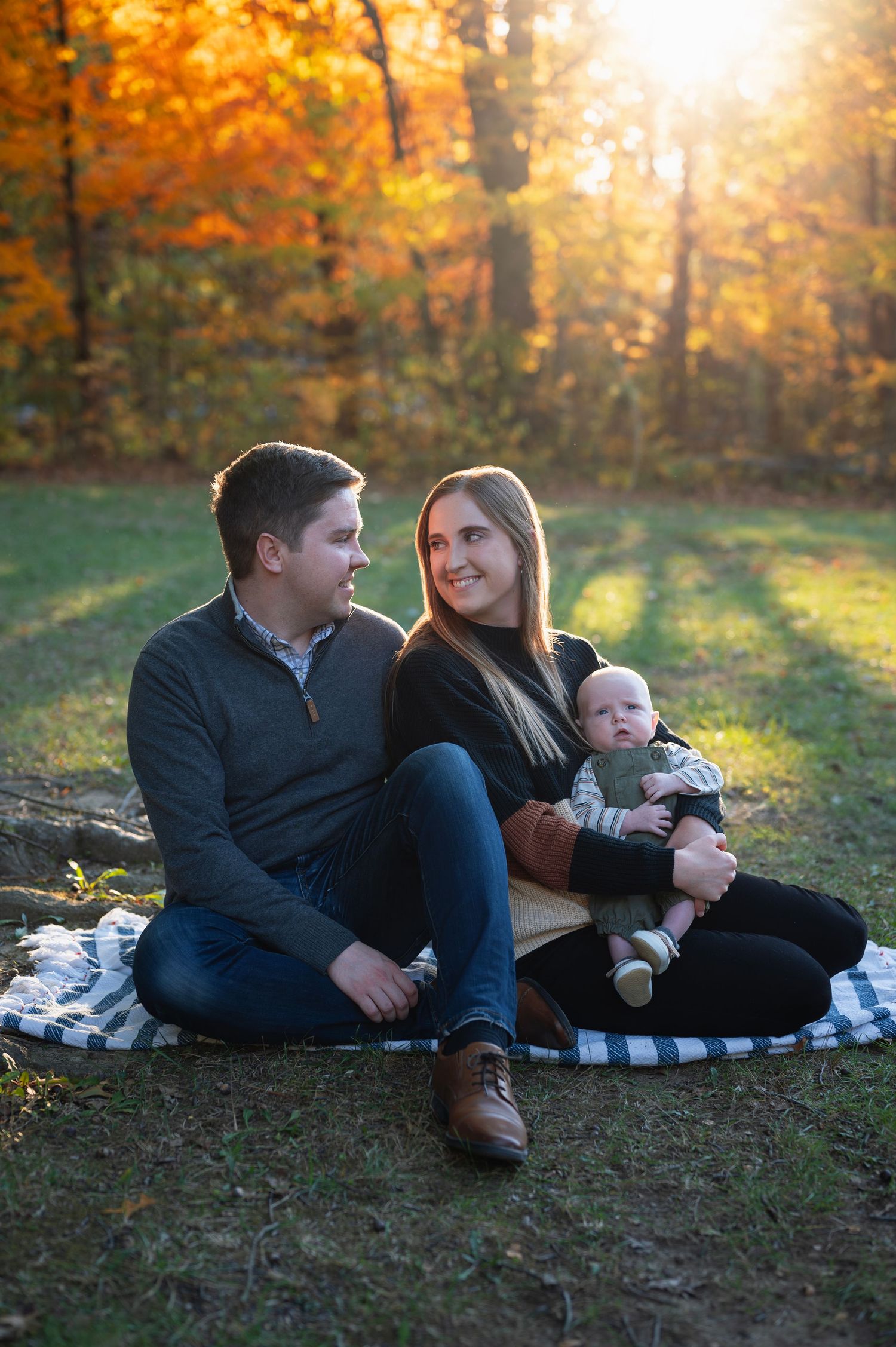 Family sitting on a blanket with sunlit golden trees behind them, smiling at each other. Highbanks Metro Park