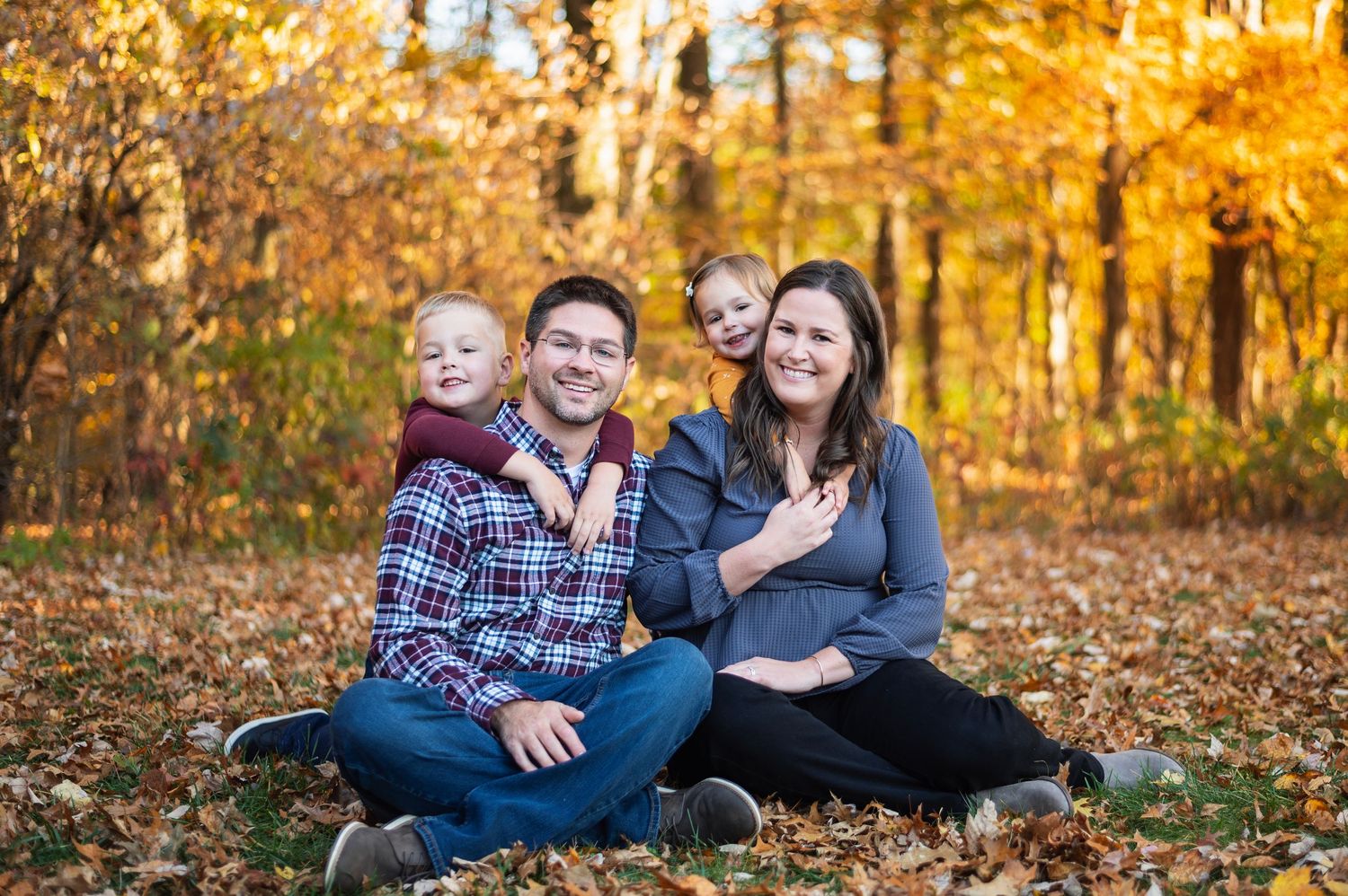 Family of four sitting in fallen leaves with vibrant fall colors in the background, children hugging their parents. Innis Woods Metro Garden