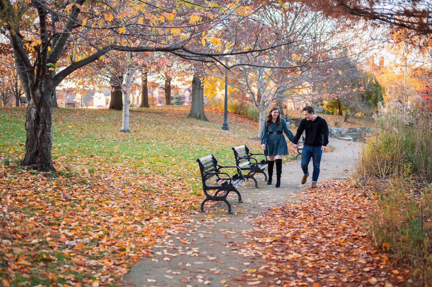 Couple walking hand-in-hand on a leaf-strewn park path lined with benches and colorful fall trees. Goodale Park