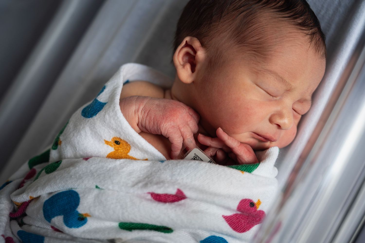 Swaddled newborn sleeps in hospital bassinet, wrapped in a colorful duck-print blanket.