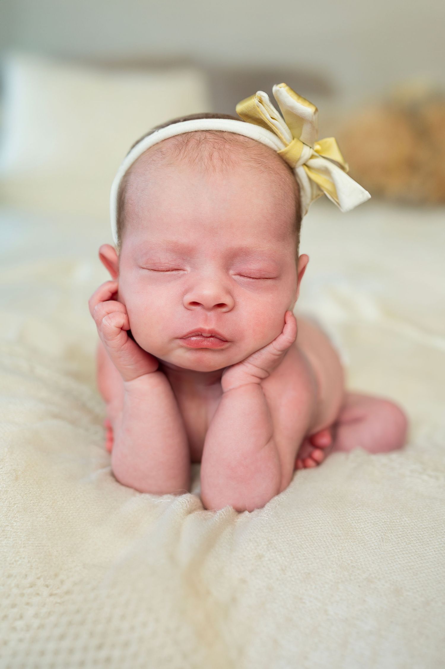 Newborn posed with hands under chin, peacefully sleeping in a classic studio setup with soft textures and a delicate bow.