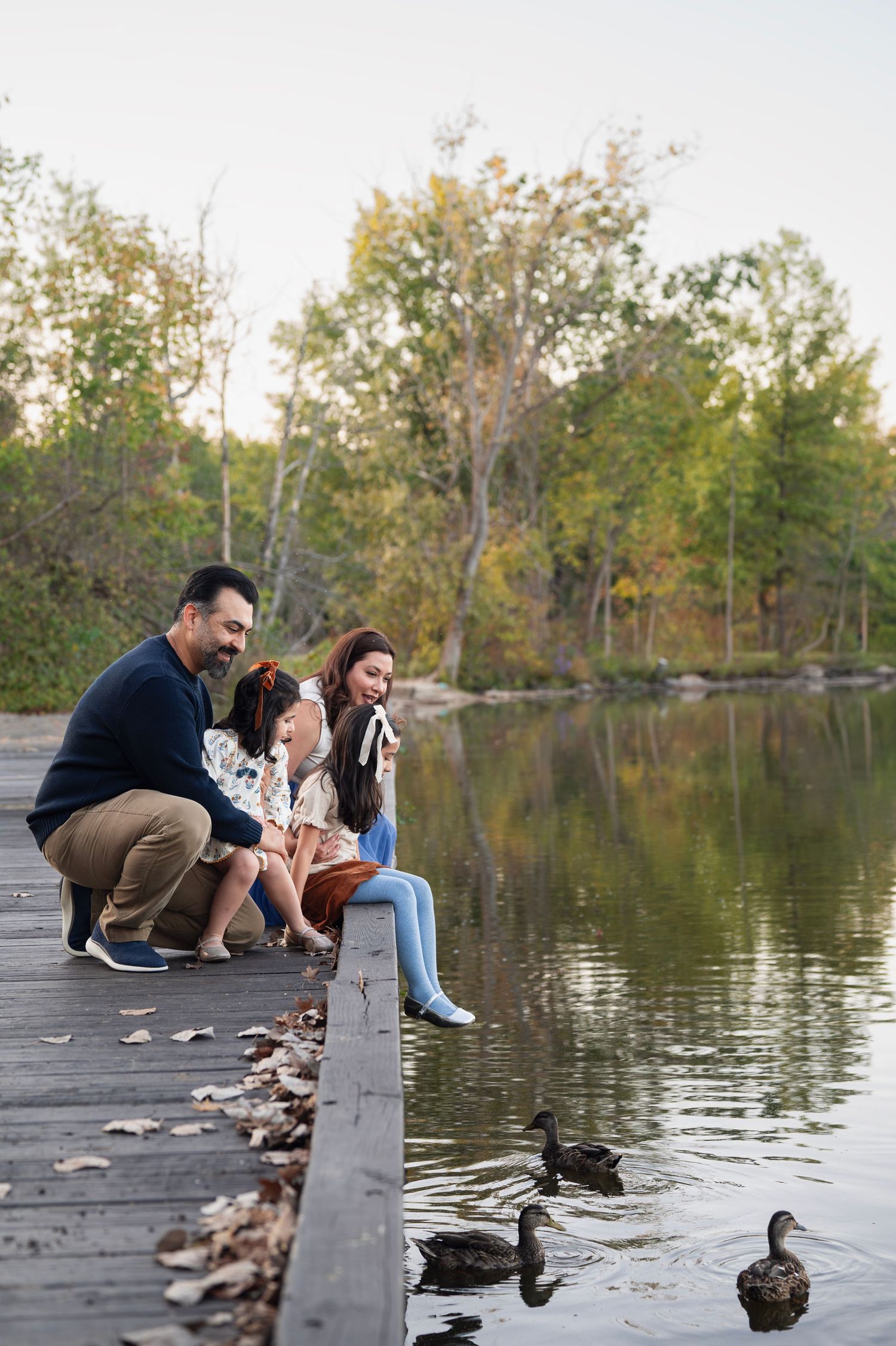 Family crouches on a wooden dock, daughters dangling their feet as they watch ducks swim in the calm lake water during fall photos.