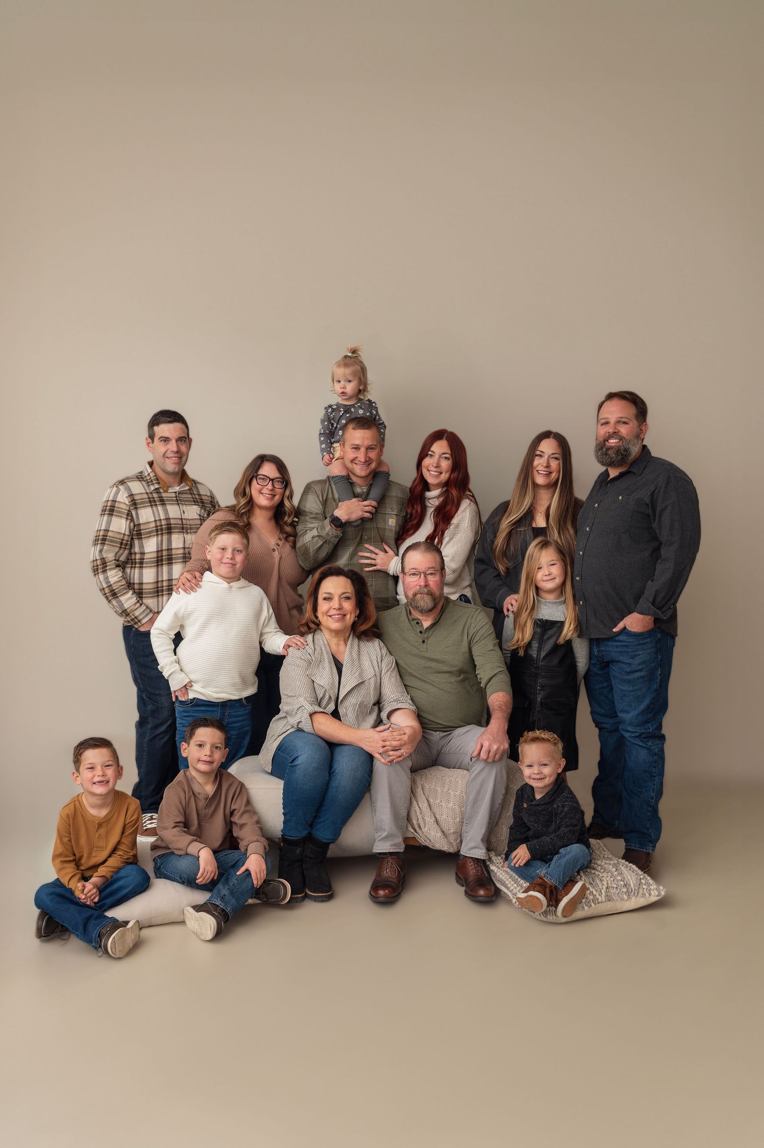 Large extended family of 14 posing in a neutral-toned studio, wearing coordinated earthy and neutral outfits.