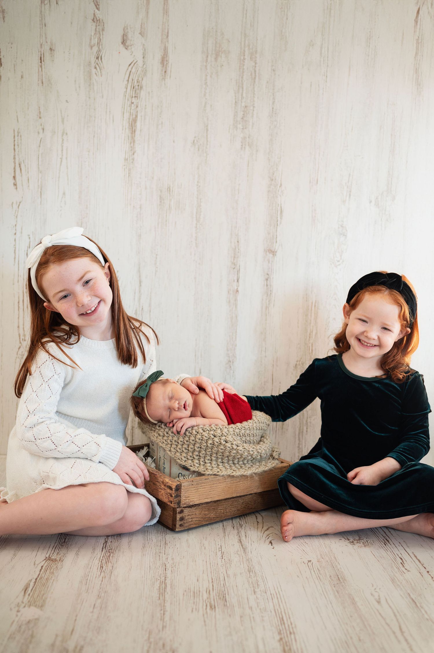 Two sisters smile next to newborn sibling in a cozy studio setup. Westerville family and newborn photography.