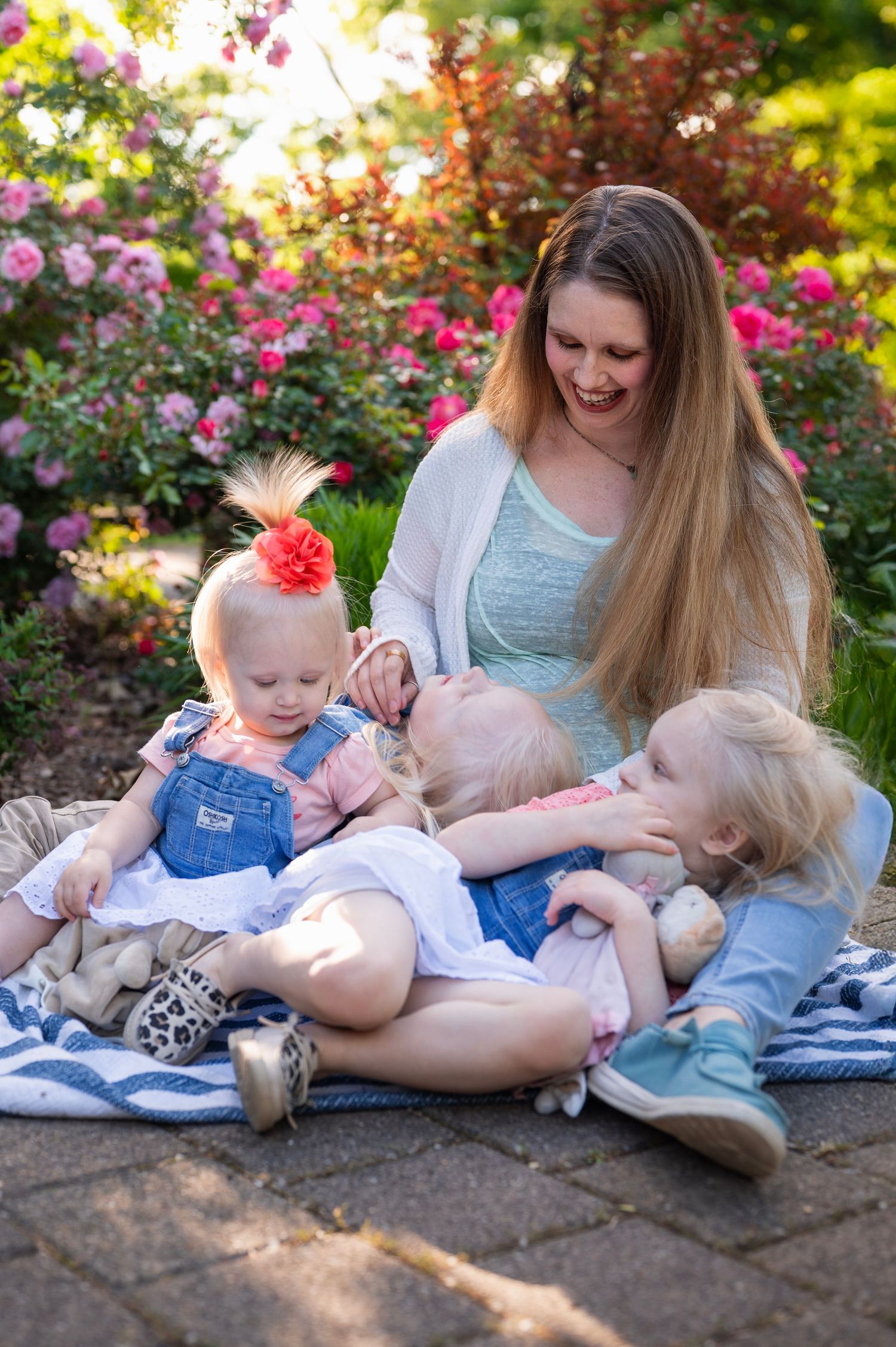 Mom laughs while cuddling her three daughters on a picnic blanket surrounded by blooming roses.