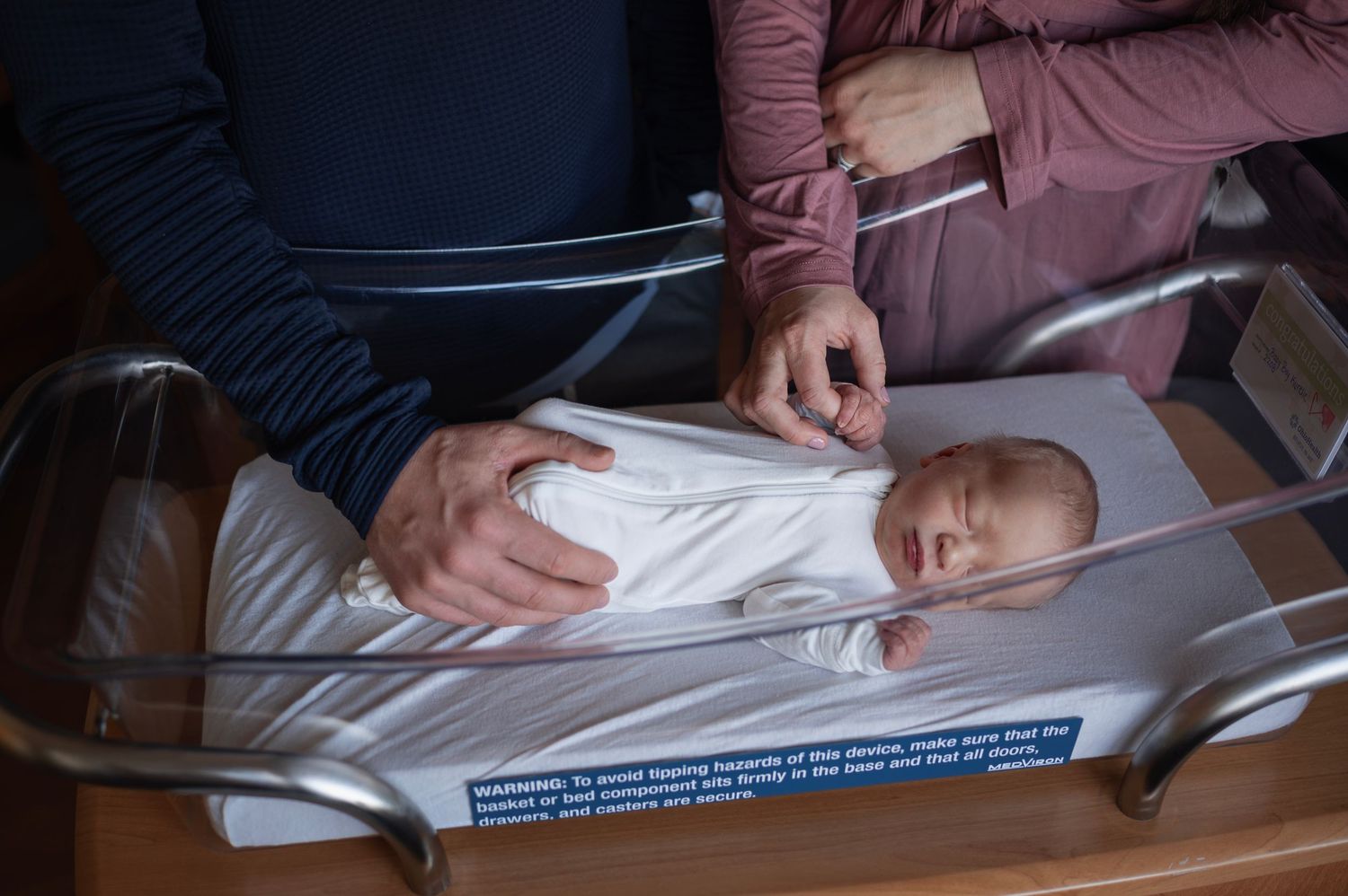 Parents gently hold baby’s hand in hospital bassinet during tender Fresh 48 session.