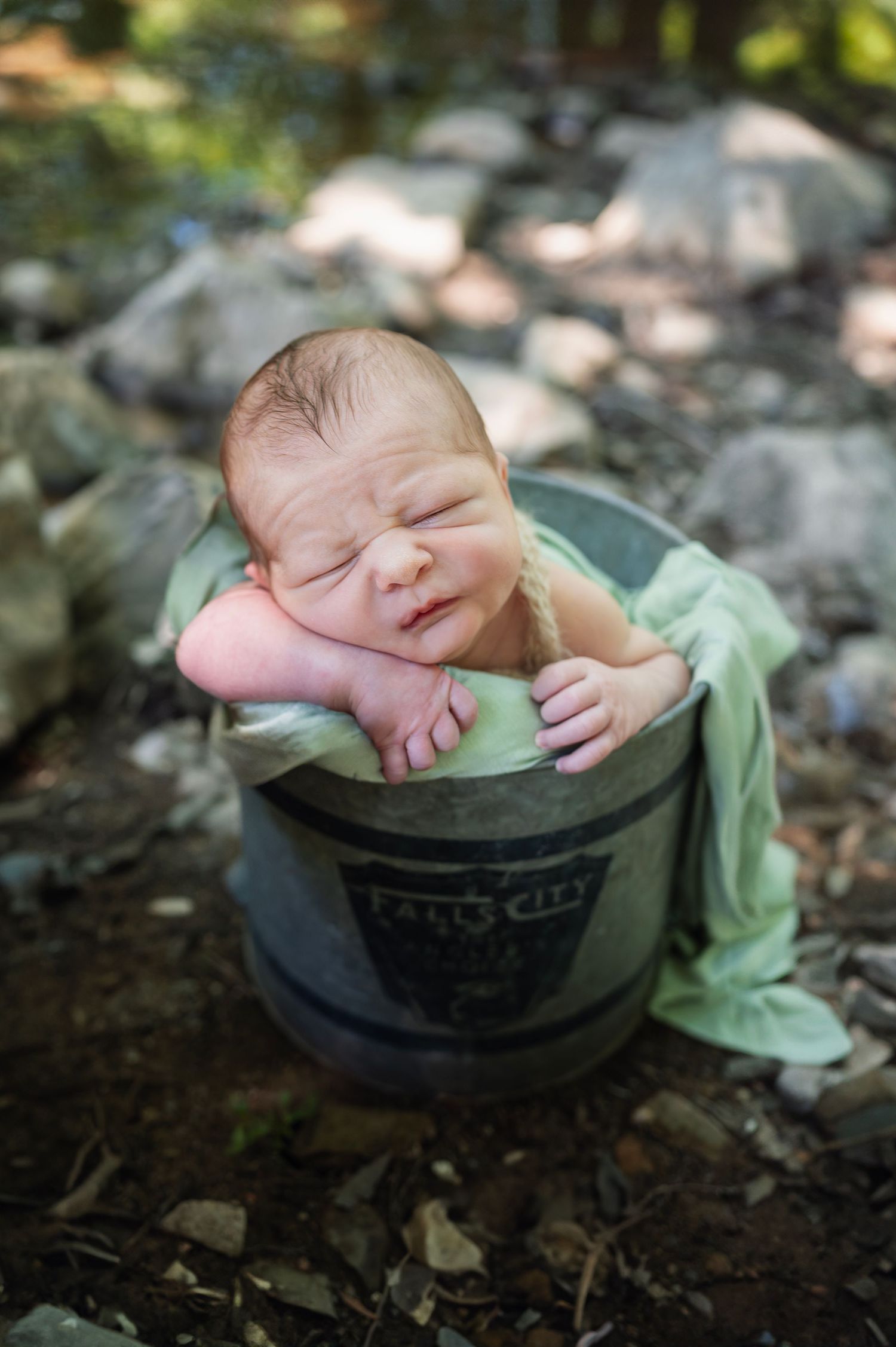 Newborn rests in a metal bucket by a rocky creek bed during an outdoor session, eyes squinting in the sunlight.