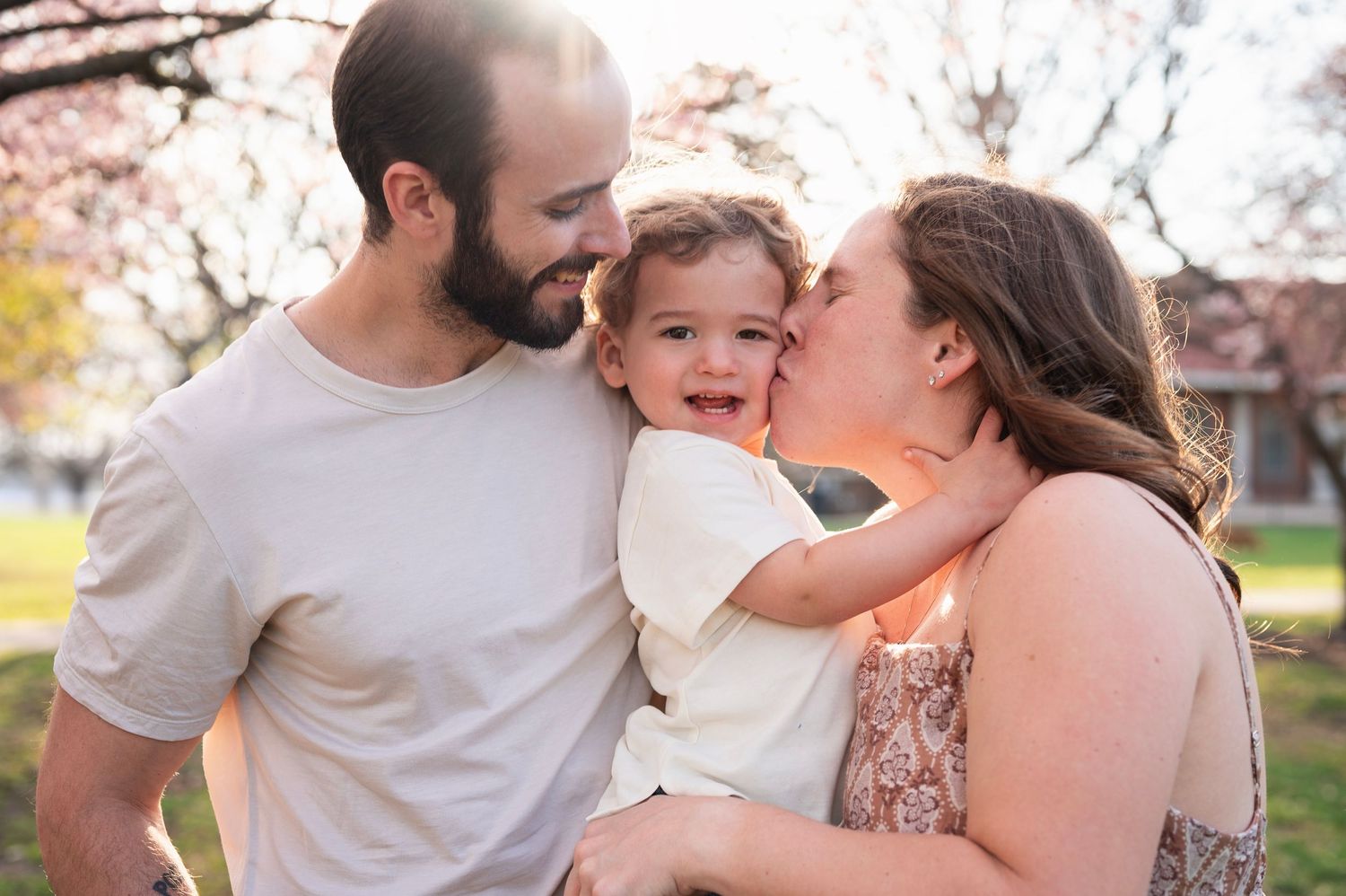 Westerville family photographer captures parents kissing their toddler during a spring session under blooming trees.