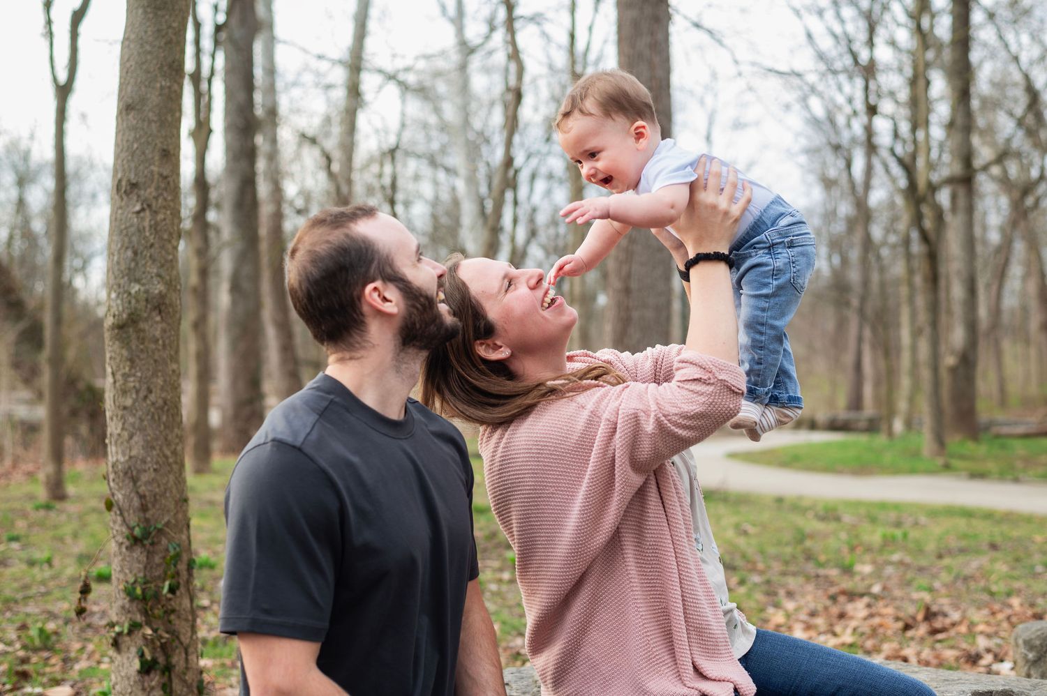 Mom lifts baby high as dad smiles in Gahanna woods. Family joy captured by Westerville family photographer.