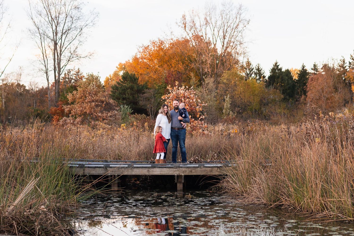 Family of four standing on a dock over a pond, surrounded by tall grasses and autumn trees reflecting in the water. Highlands Aquatic Center