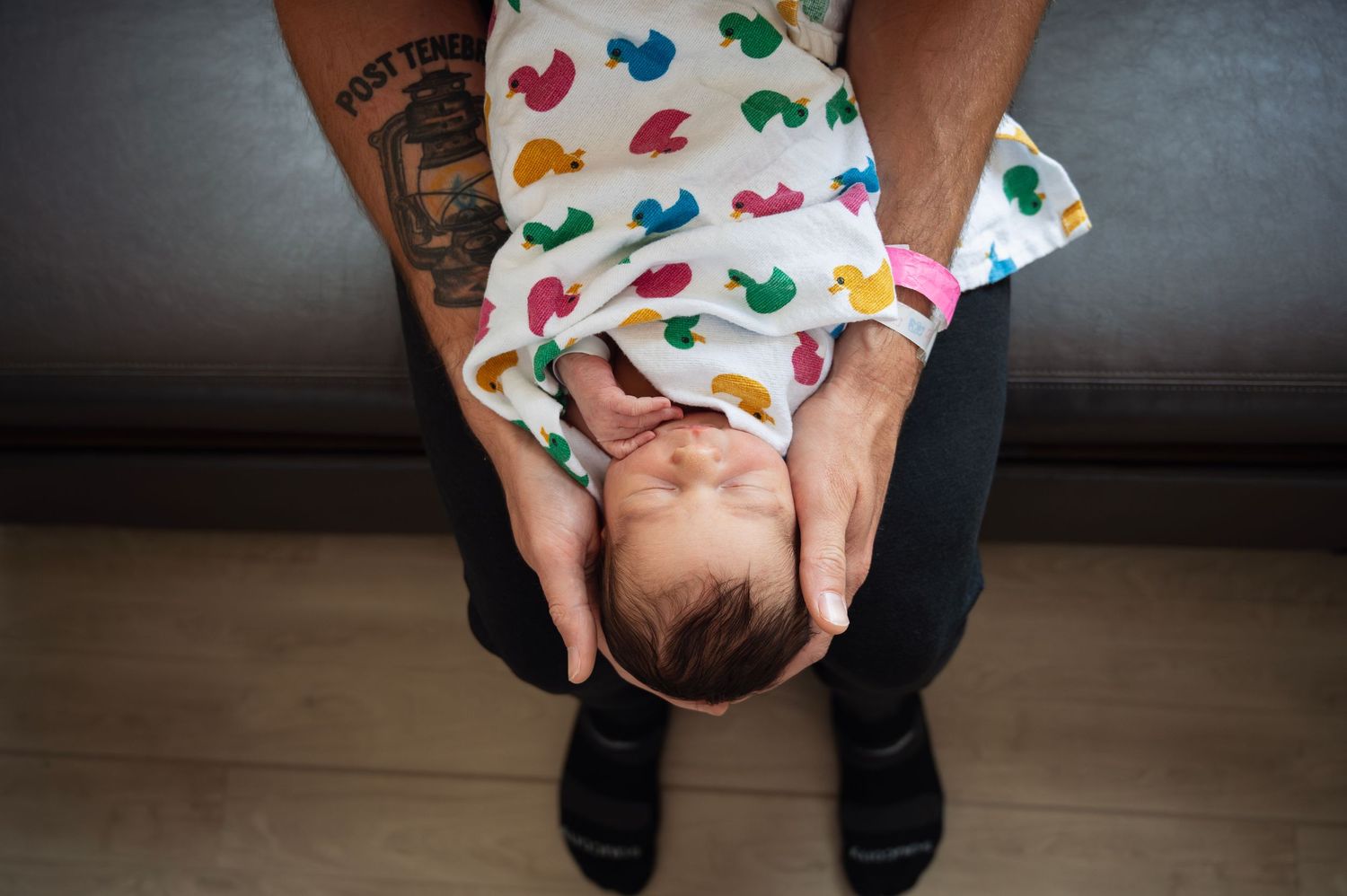 Tattooed dad cradles newborn wrapped in colorful duck swaddle, hands gently supporting baby’s head.