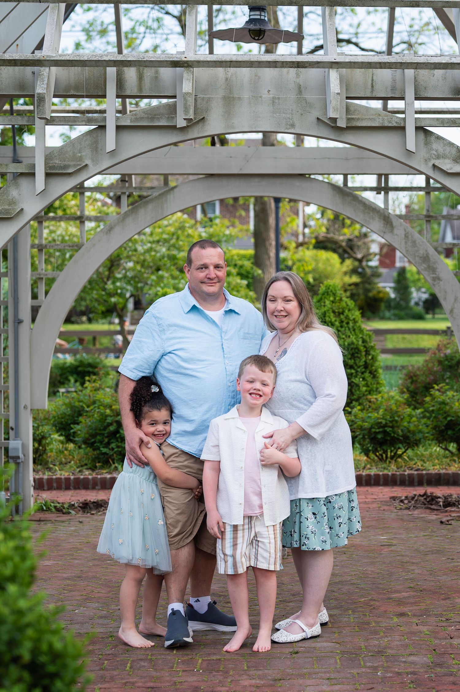 Family of four standing under a wooden garden arch, smiling while wearing pastel and neutral tones.