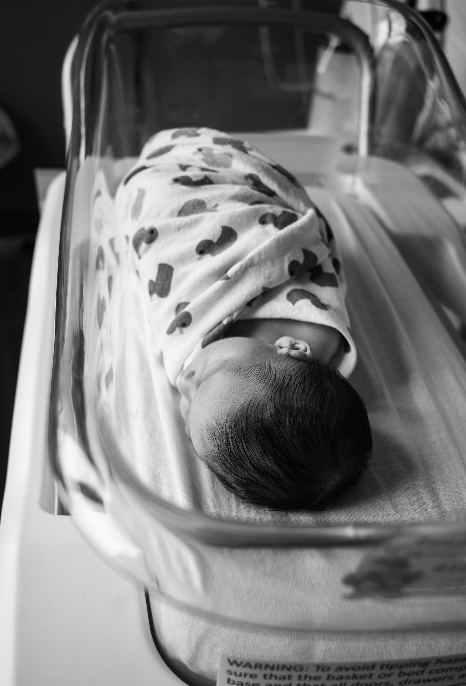 Black and white Fresh 48 hospital photo of a newborn swaddled in duck print, resting in a bassinet after birth.