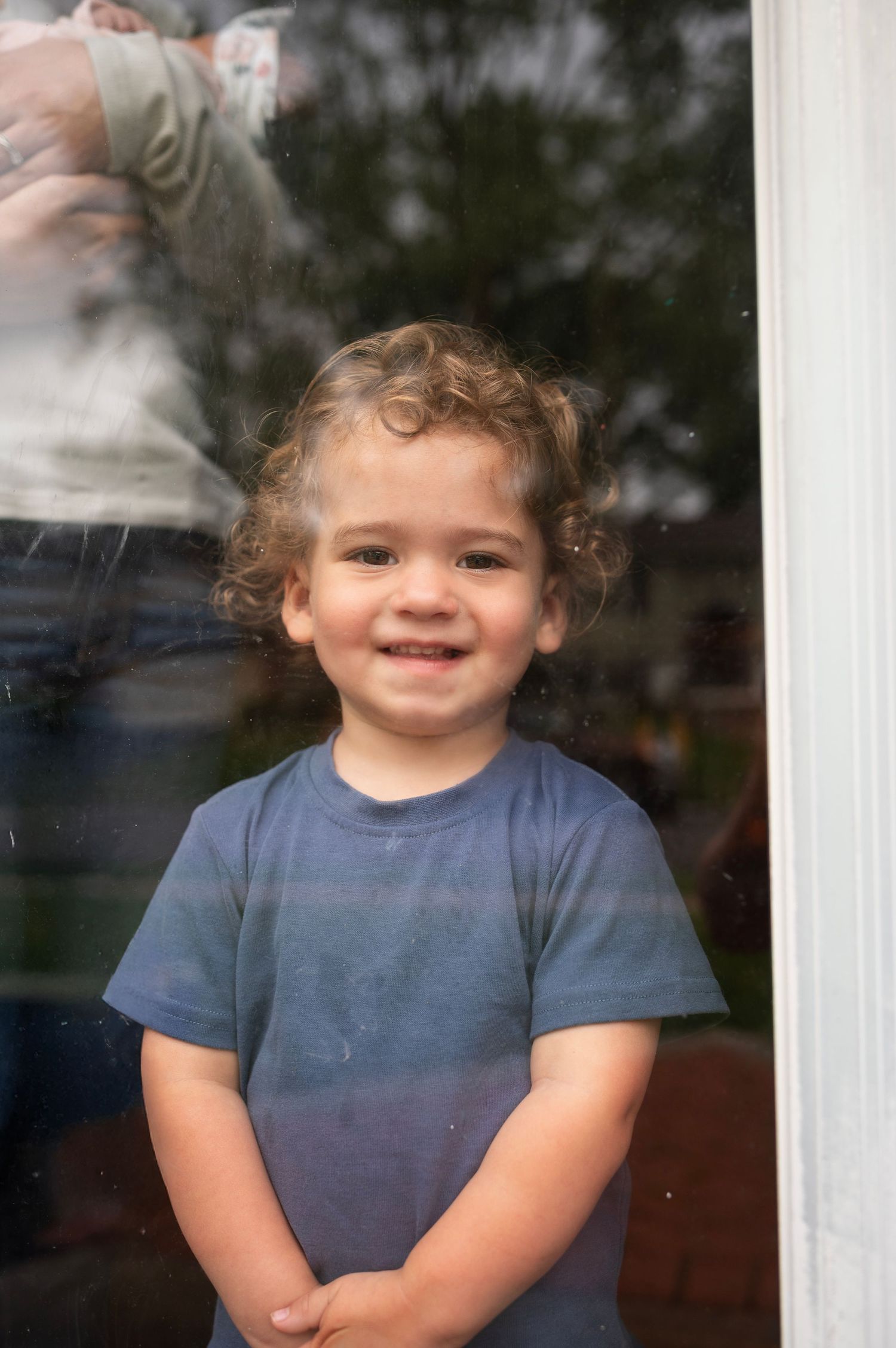Toddler smiles through a window while baby sibling is cradled behind him during an in-home lifestyle session.