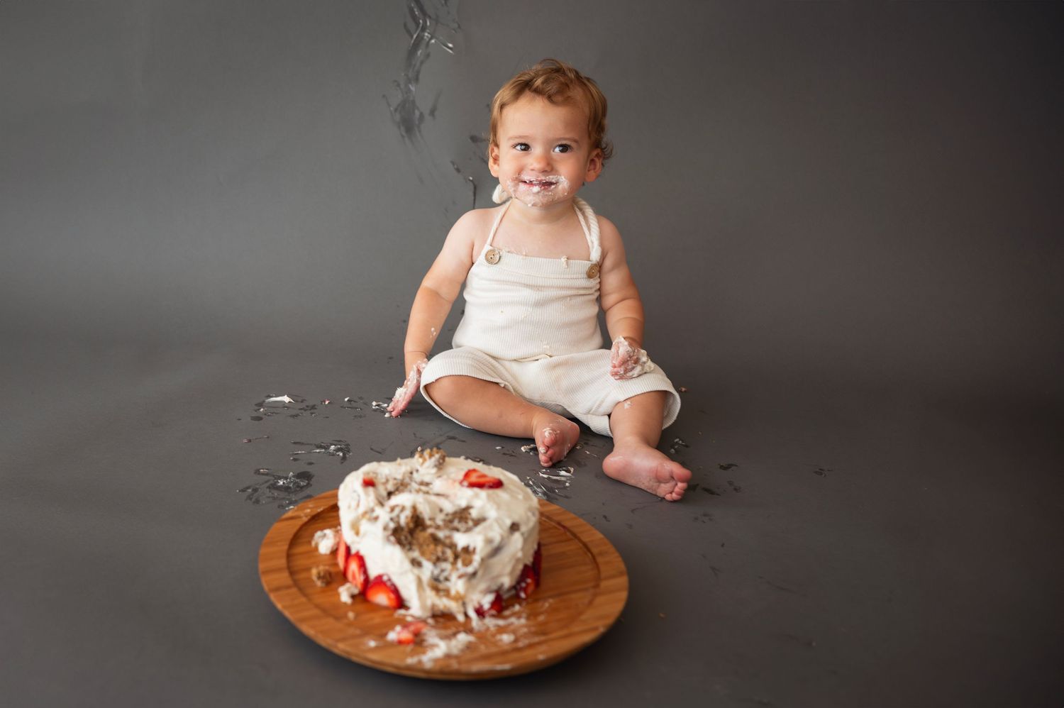 One-year-old grins after smashing a strawberry cake, wearing a cream romper with frosting on hands and face.