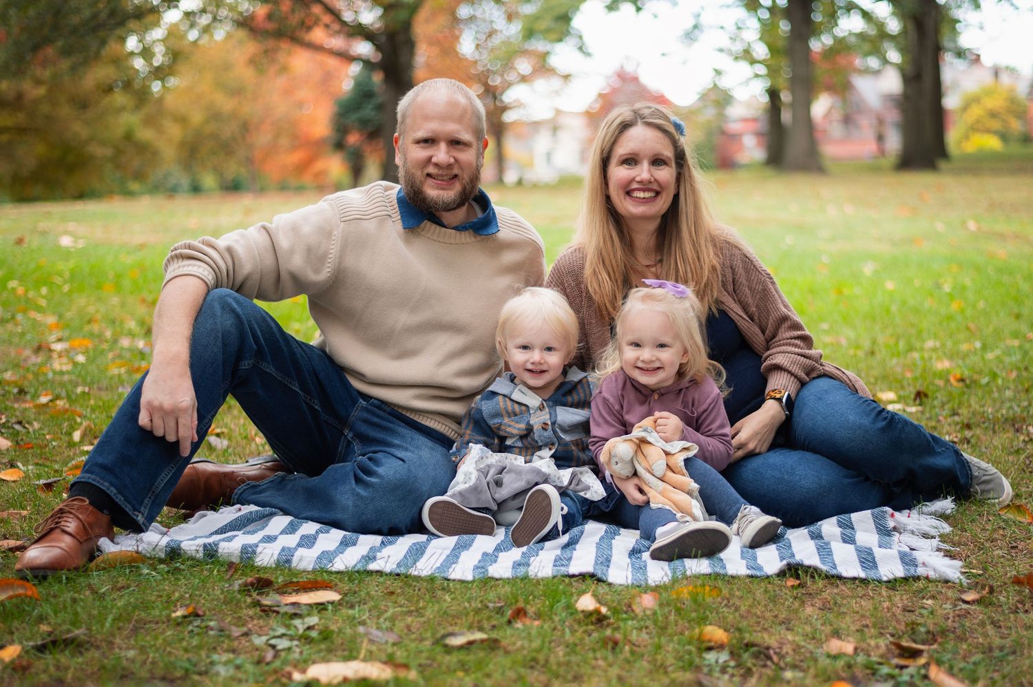 Family of four sitting on a blanket in a park with fall colors in the distance, smiling warmly. Goodale Park