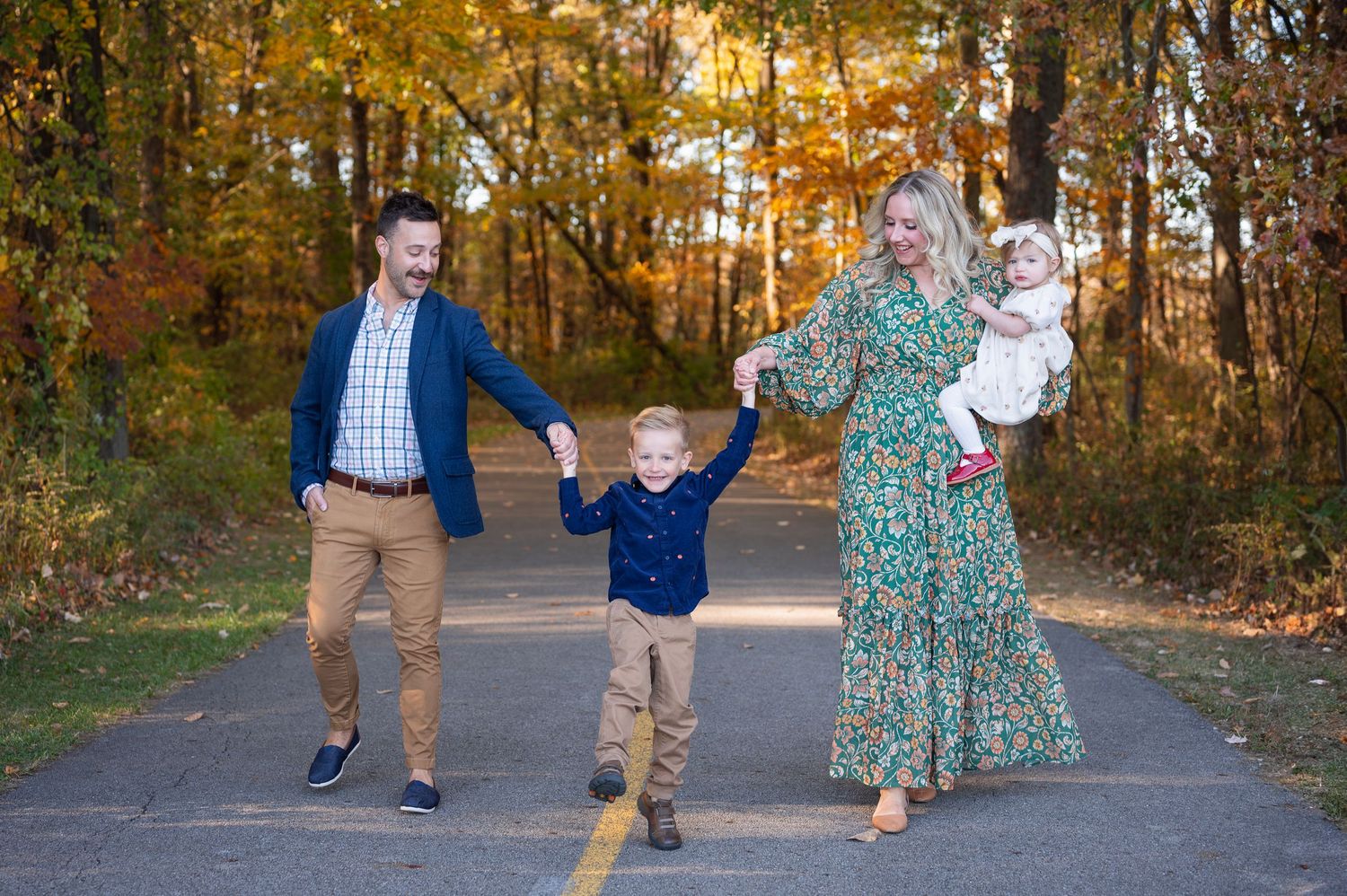 Family of four walking hand-in-hand down a wooded path in fall, wearing coordinated blue, green, and neutral tones.