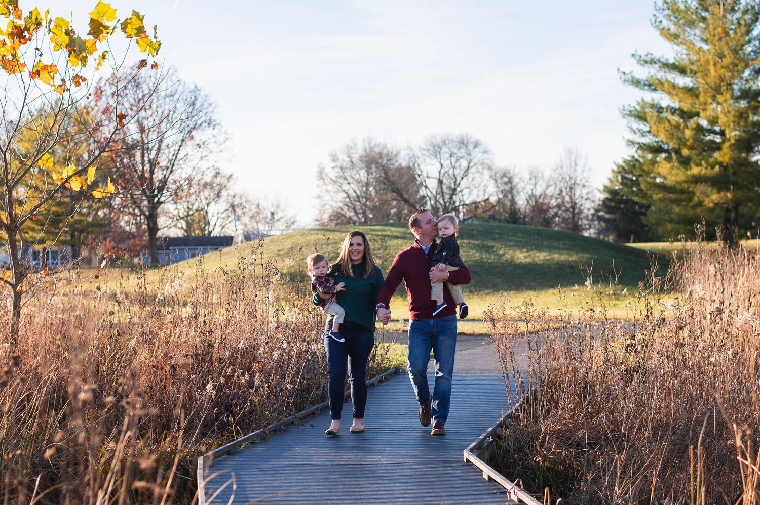 Family of four walking on a boardwalk through tall grasses with late fall foliage in the background. Highlands Aquatic Center