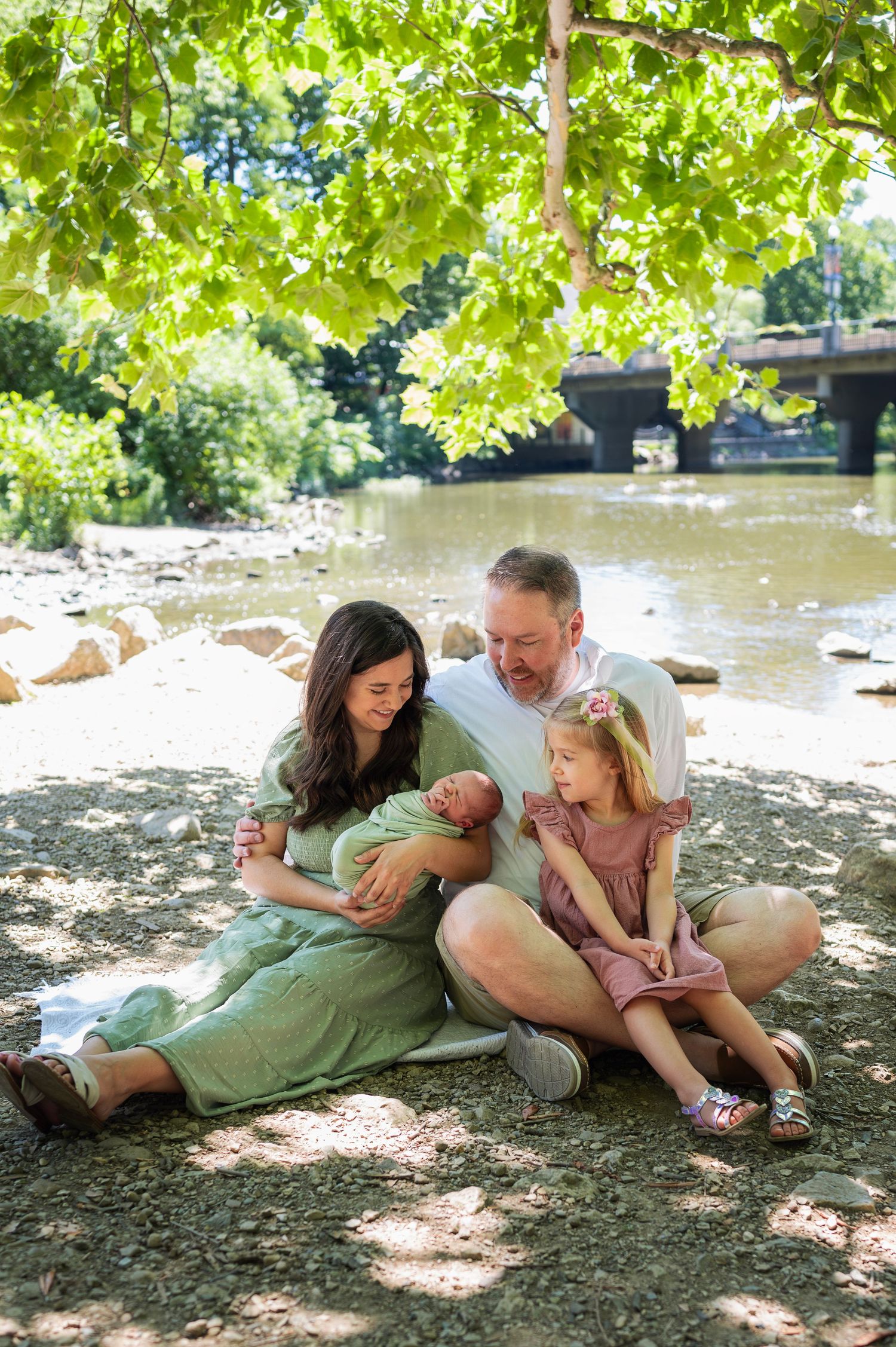 Family of four sits under a tree by the river, snuggling their newborn while big sister beams at the baby.
