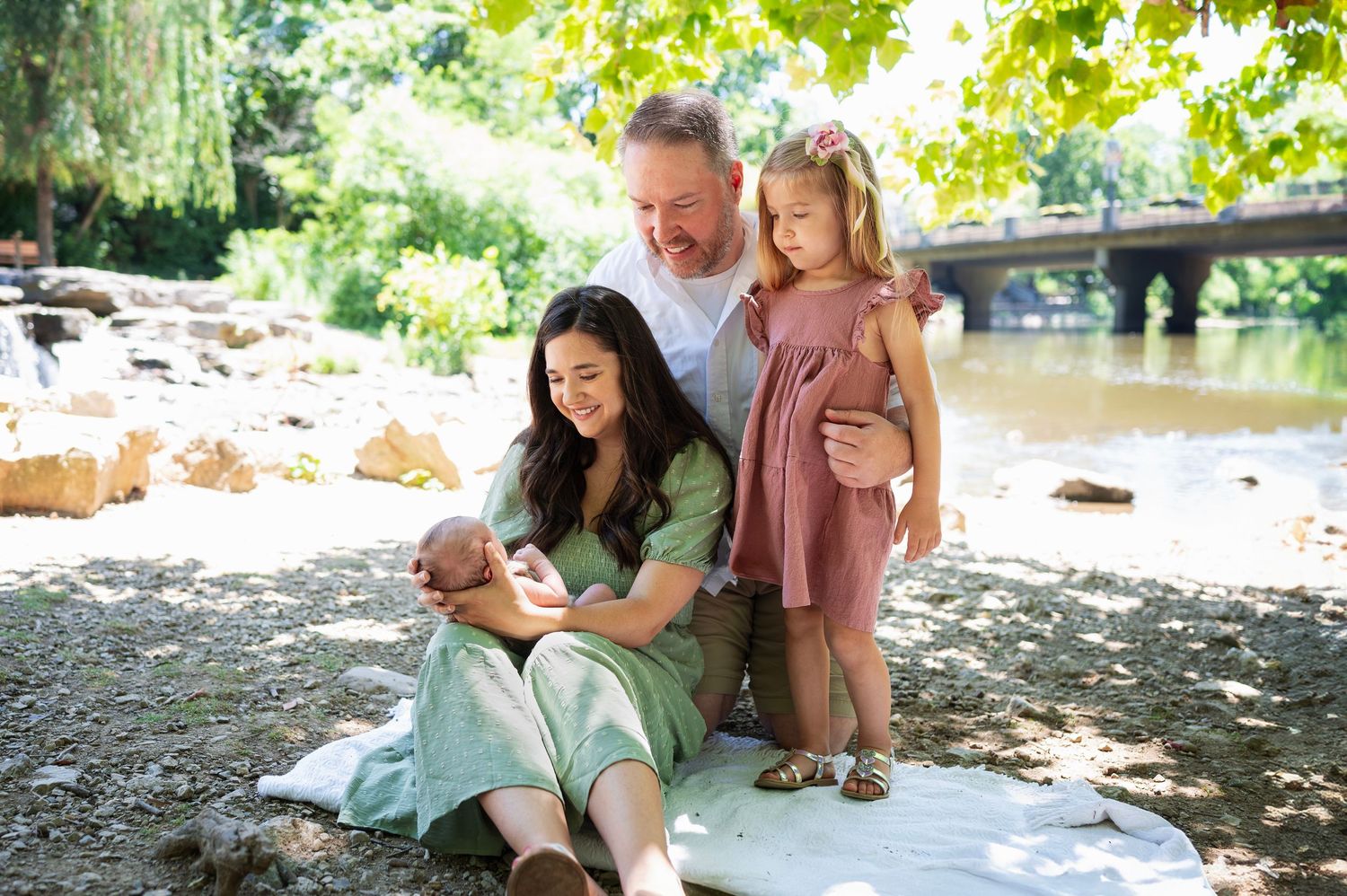Family sitting by a river under dappled sunlight, mom in a sage green dress cradling newborn, dad and older daughter in soft pink