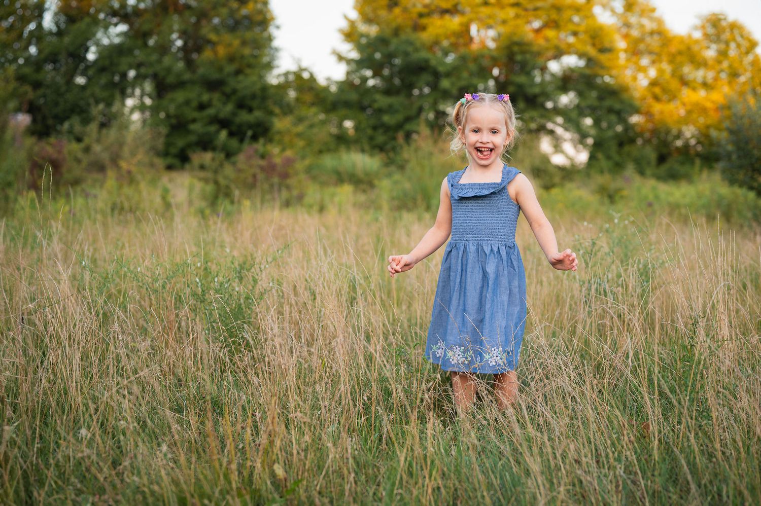Girl laughs in tall grass at golden hour. Candid child portrait by Westerville family photographer.