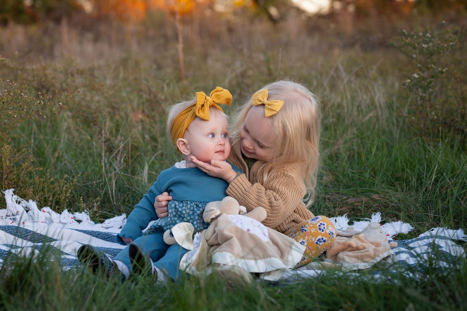 Big sister gently holds her baby sister’s face as they sit on a blanket in a golden fall field.