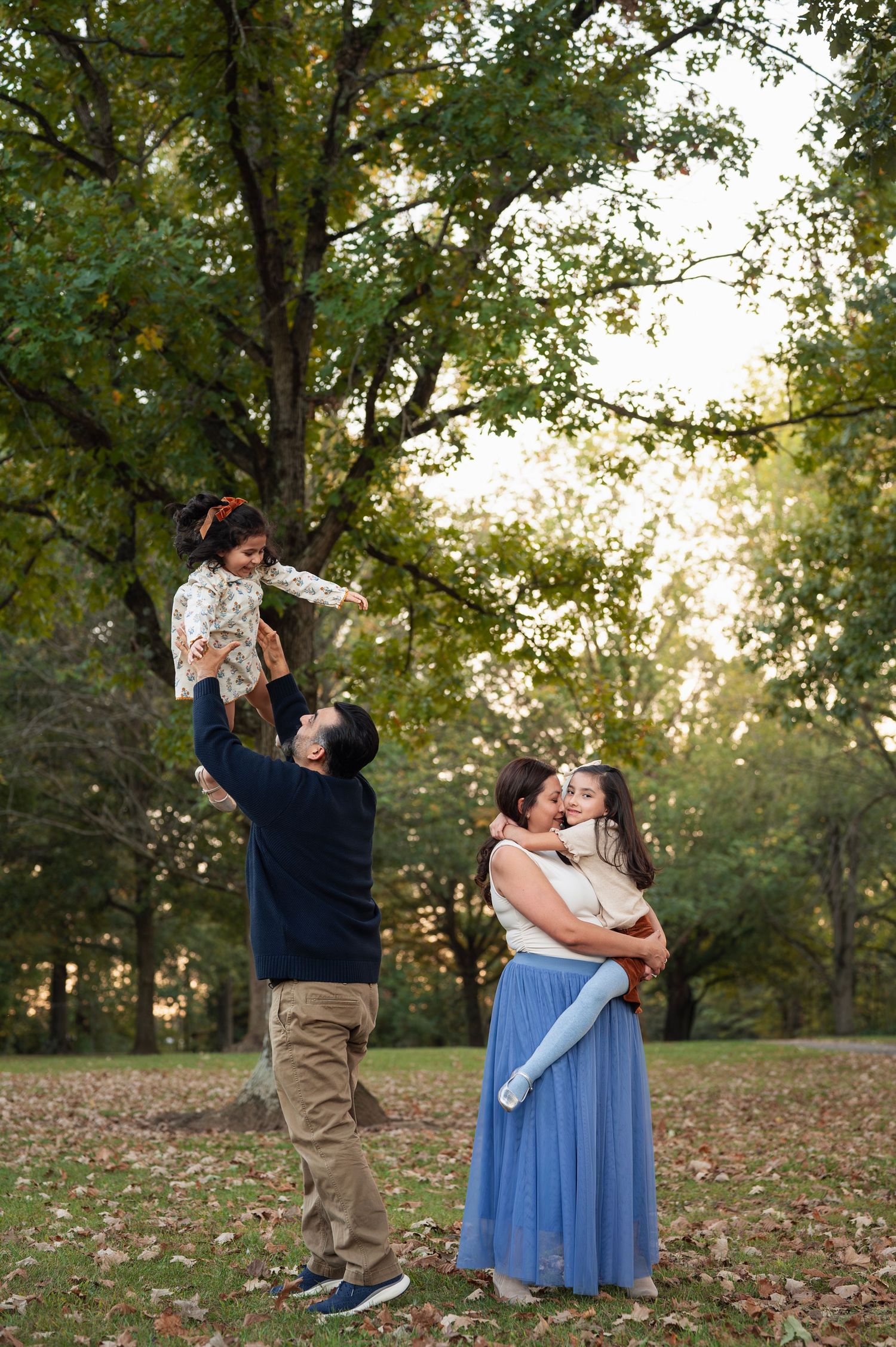 Dad lifts his laughing toddler high while mom snuggles their older daughter, framed by tall green trees and crunchy fall leaves.