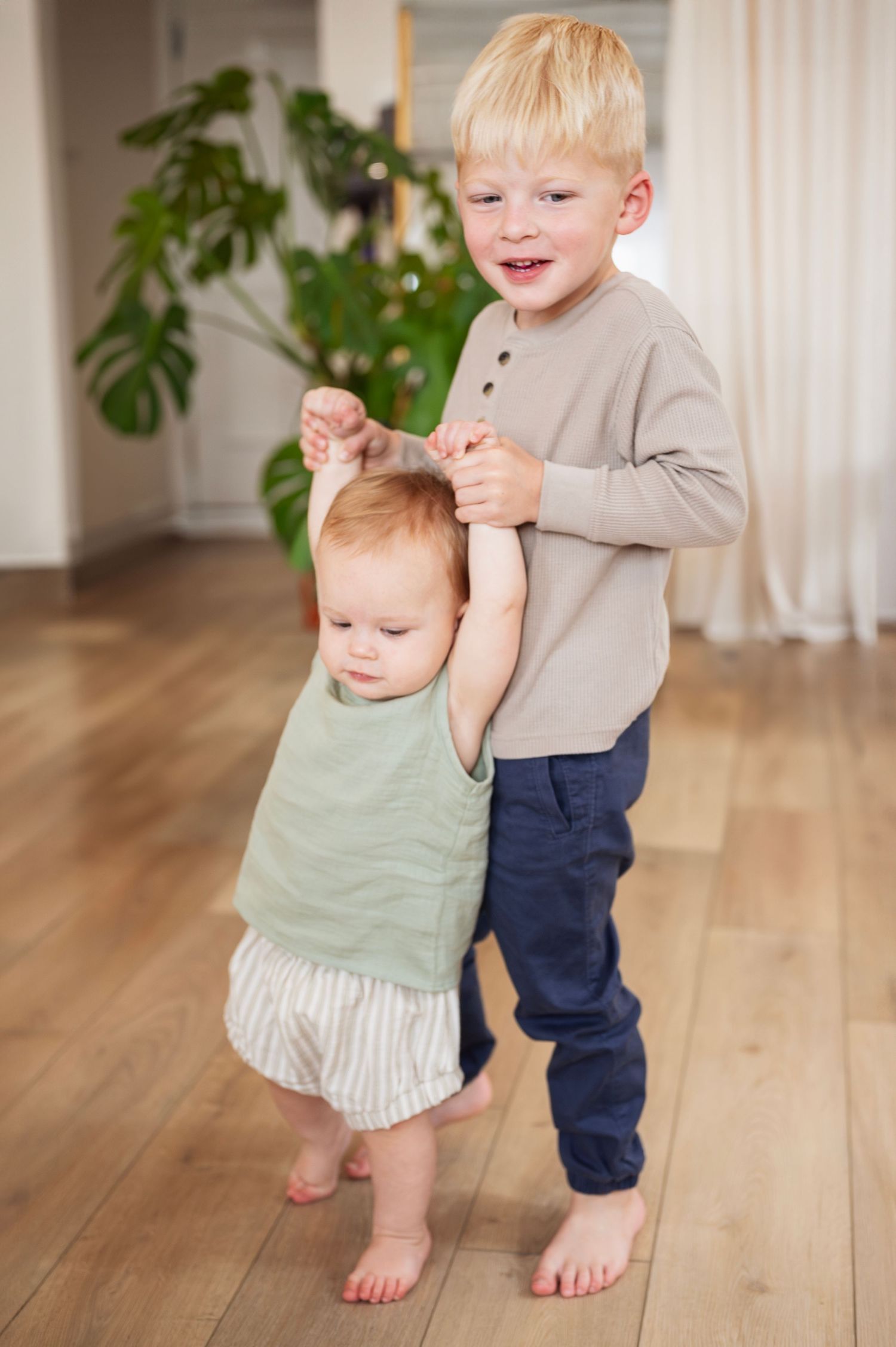 Big brother holds baby sister’s hands and helps her walk barefoot during an in-home milestone session.