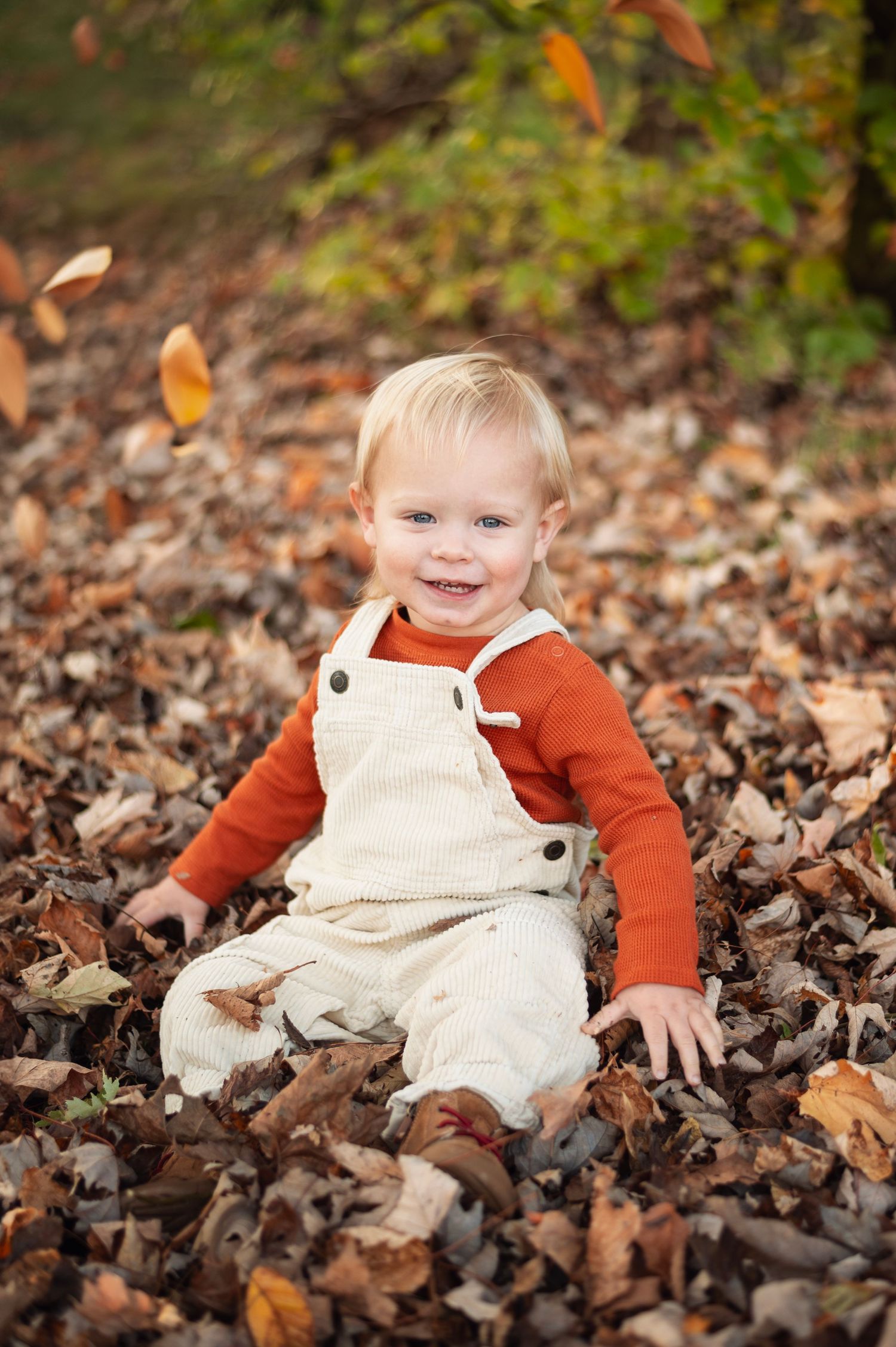 Toddler in cream overalls and an orange sweater sitting in fallen leaves, smiling at the camera.