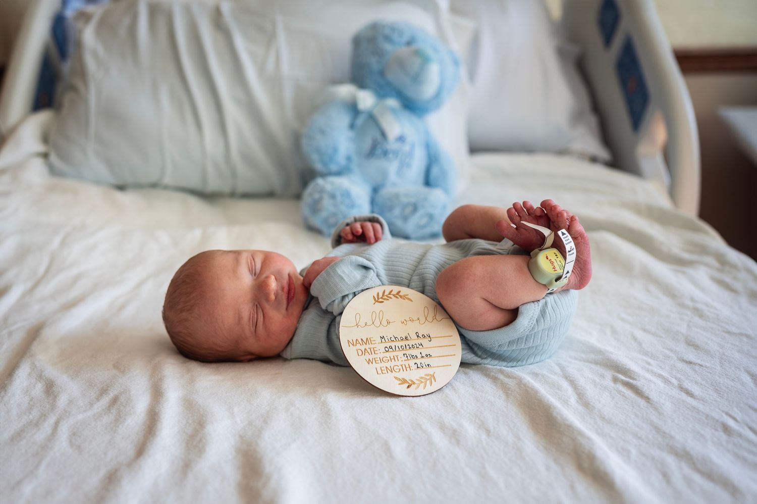Newborn boy rests on hospital bed with a blue teddy bear and wooden name sign showing birth details.