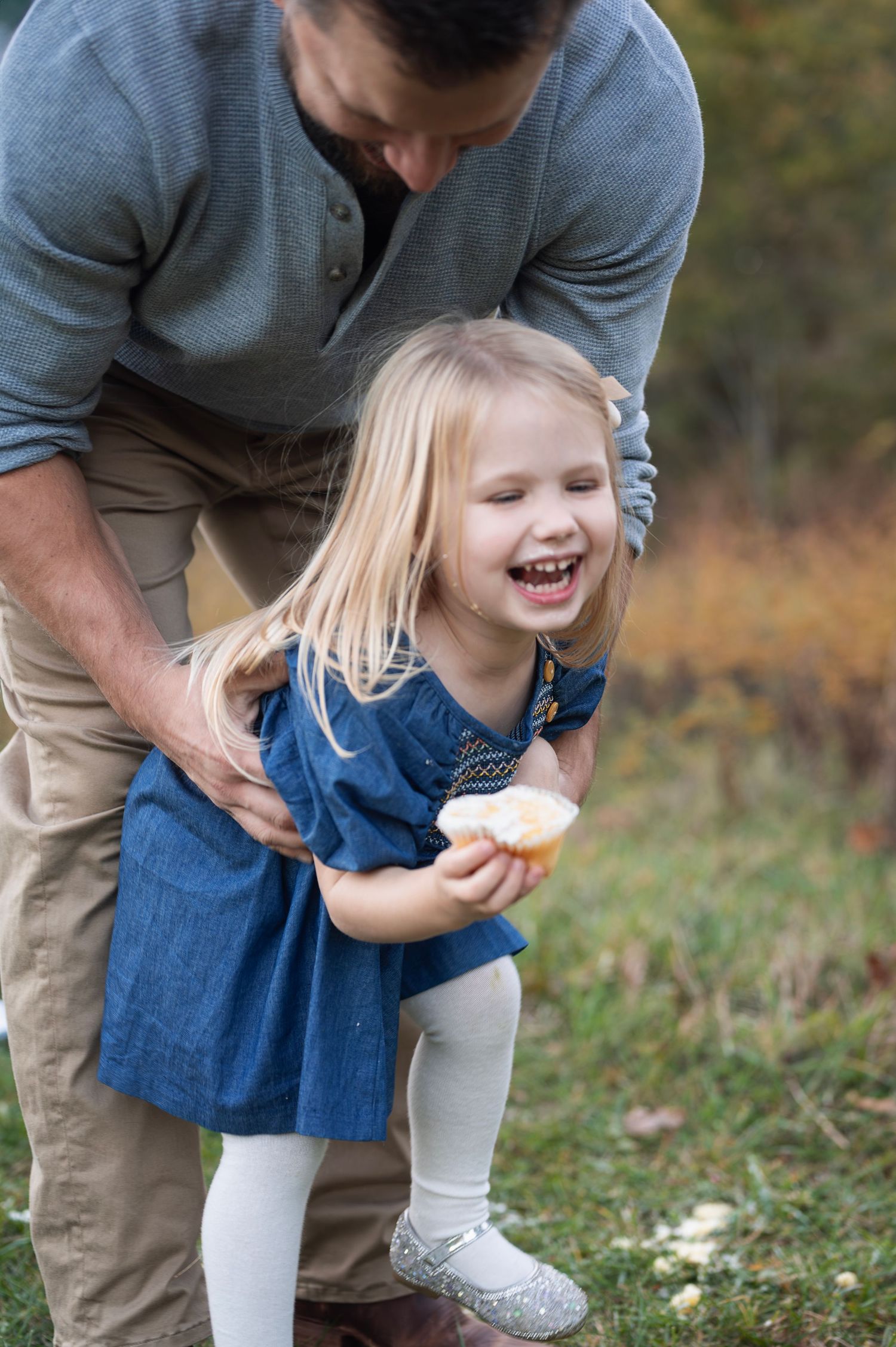 Little girl giggles with a cupcake in hand as dad scoops her up during a joyful outdoor session.